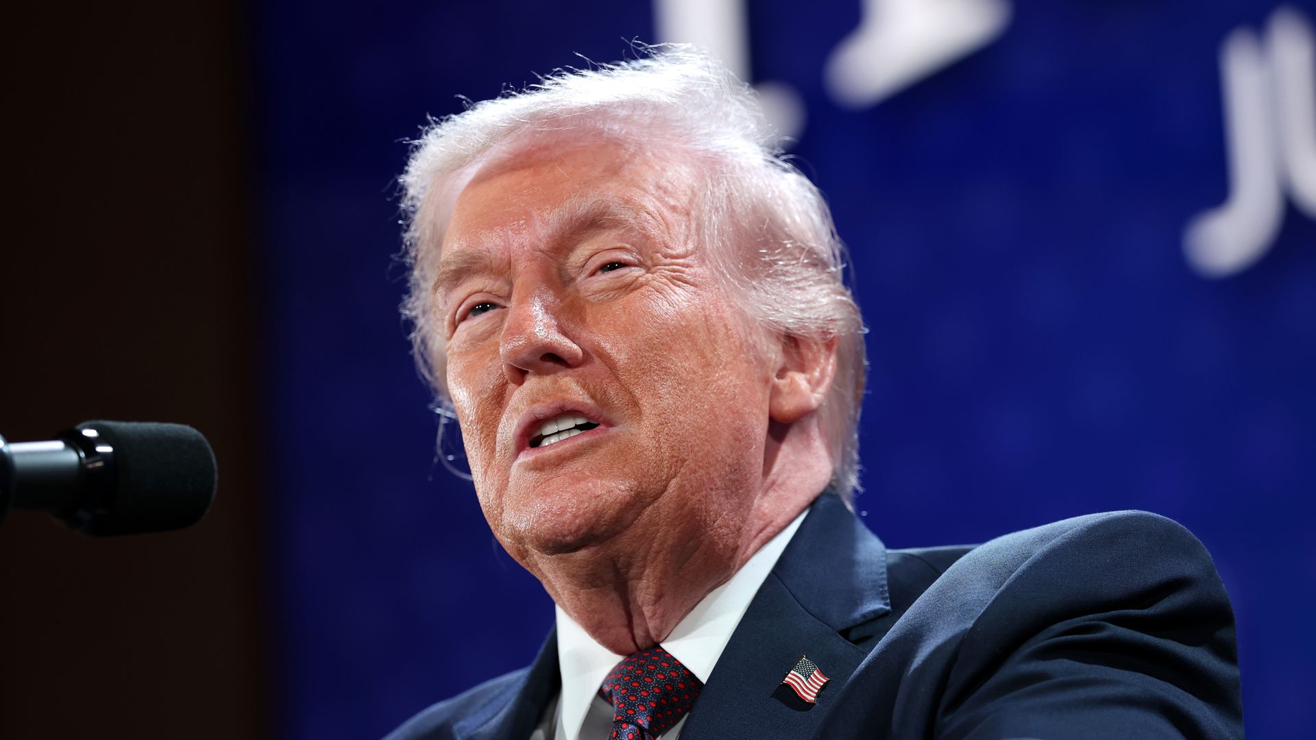 Trump speaking into a microphone wearing a blue suit jacket with an American flag pin, white collared shirt and red and blue patterned tie