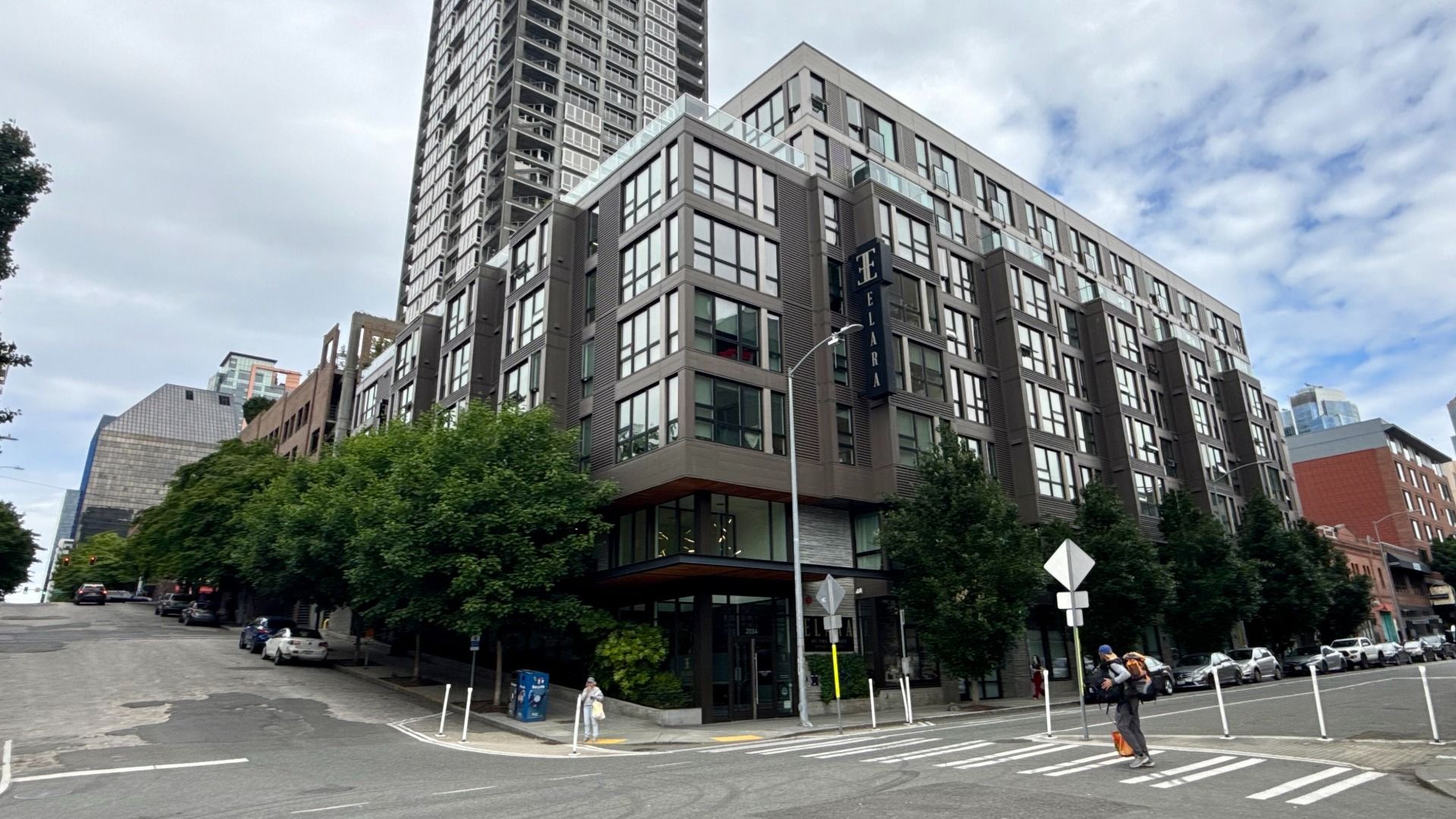 Urban street scene with a modern gray, multi-story apartment building at the corner and a tall tower behind it. Trees line the sidewalk, crosswalk markings, parked cars, and pedestrians nearby.