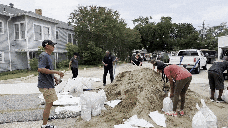 Several people fill white sandbags from a large sand pile in a residential area with gray houses and trees, likely preparing for flooding or a storm. Vehicles are parked nearby.