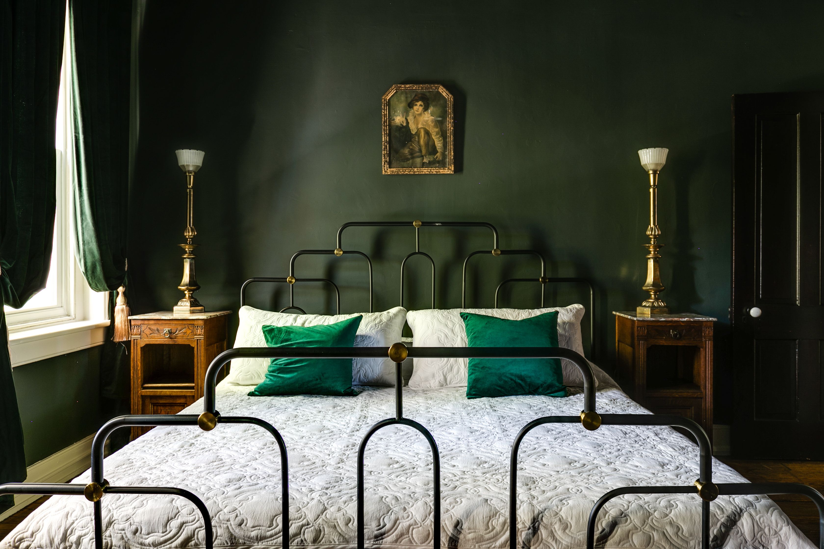 A bedroom with dark green walls, a creepy framed pic, macabre-looking lamps and a bed with a white comforter and green throw pillows.