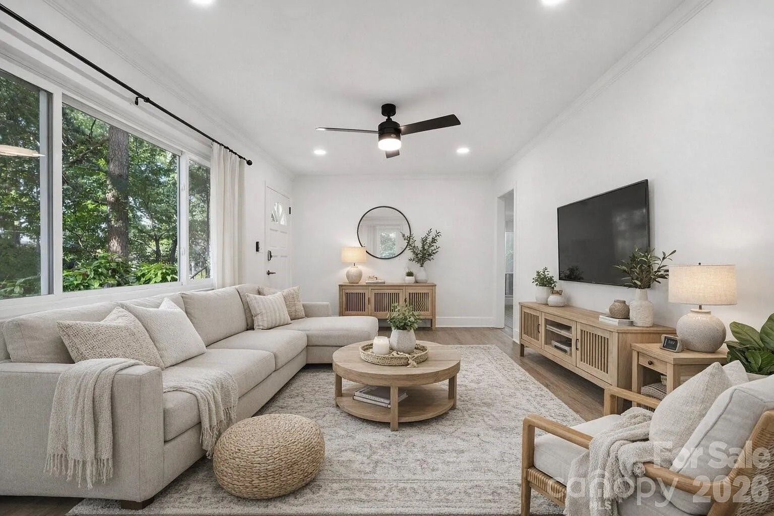 Bright living room with large windows showing green trees, beige sectional sofa with blankets and pillows, round wooden coffee table, woven pouf, wooden TV stand, and neutral decor.