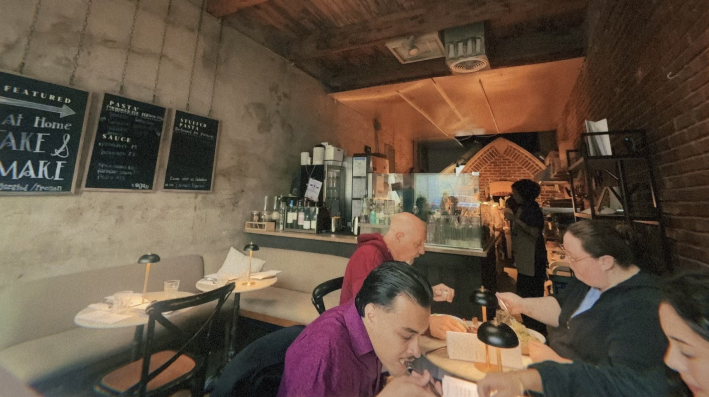 Cozy brick-walled cafe with chalkboard menus on the left, beige booth seating, and round tables with small lamps. Four diners at a table, including a bald man in a magenta shirt near the open kitchen and brick oven.