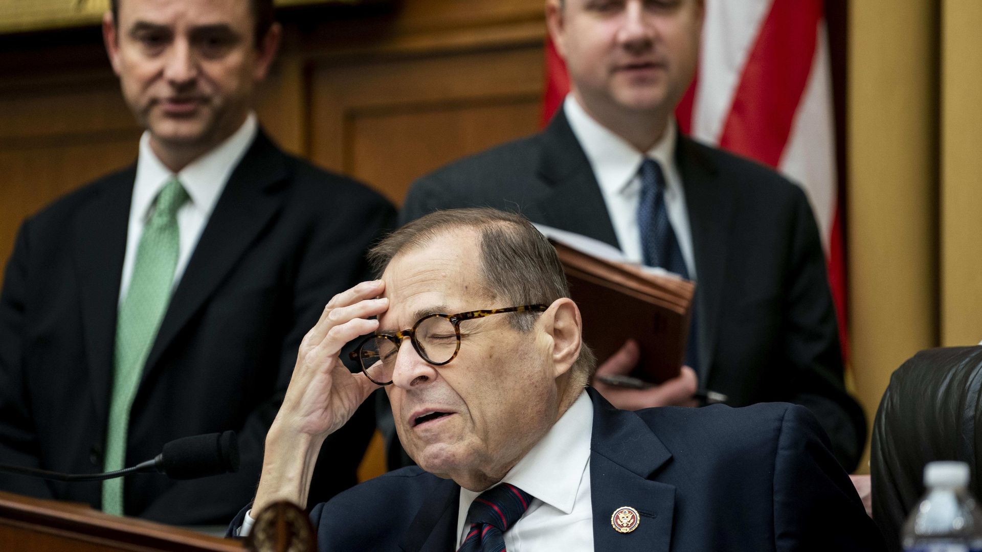 In this image, Nadler shuts his eyes and holds his head while sitting down. Two men stand behind him, on either side of him.