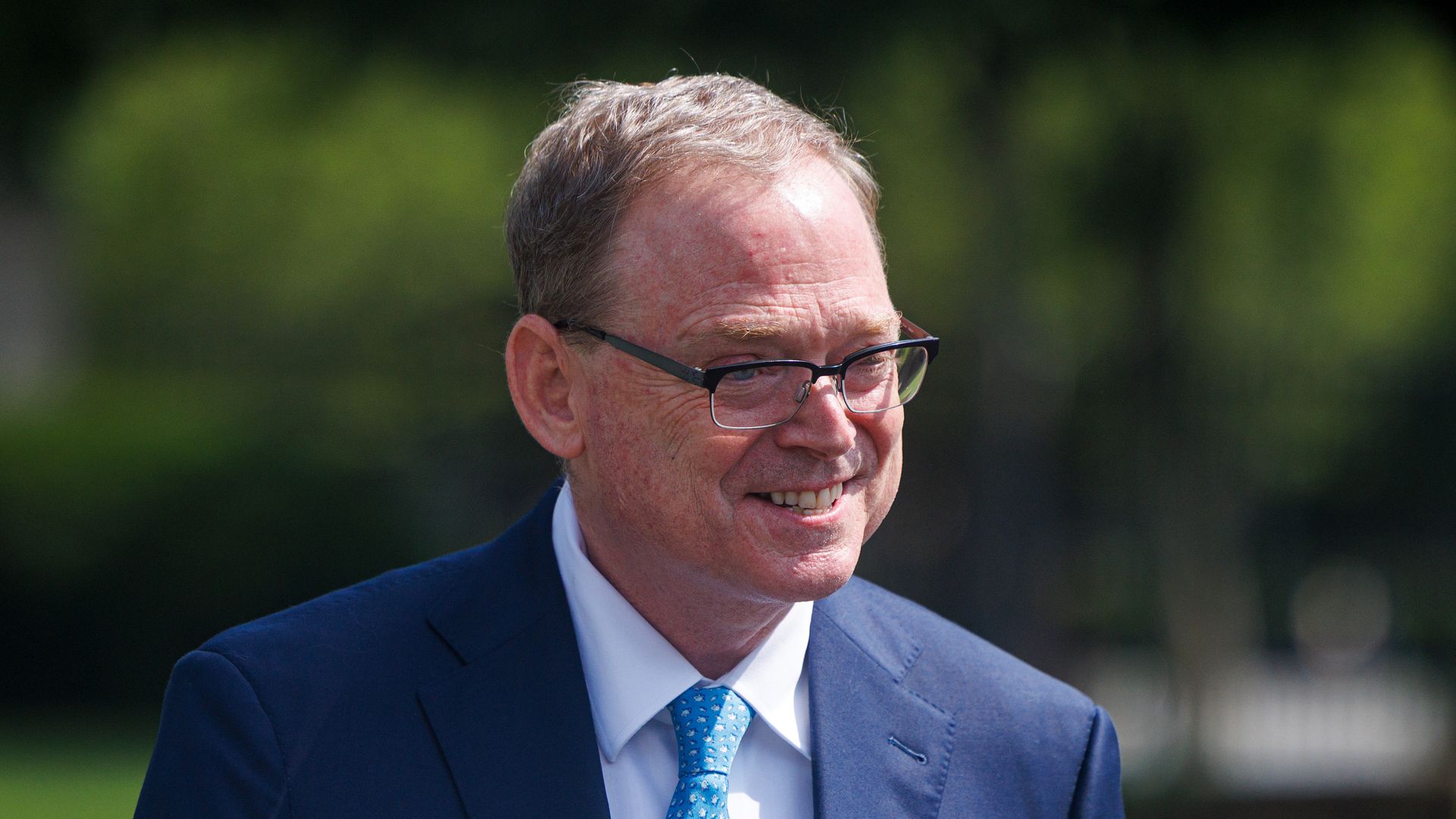 Smiling middle-aged man wearing black glasses, a dark blue suit, white shirt, and light blue patterned tie, outdoors with blurred green background.