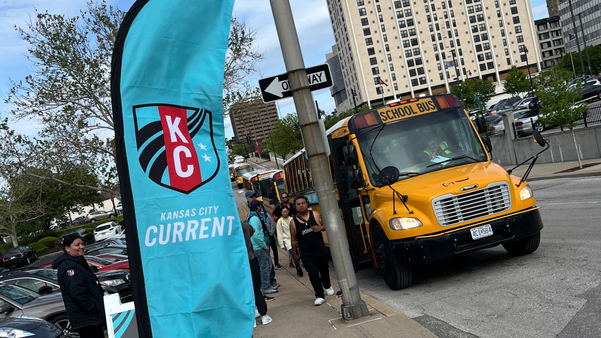 Yellow school bus on a city street with pedestrians along the sidewalk and a tall beige building in the background. A bright blue Kansas City Current banner with the KC emblem dominates the left.