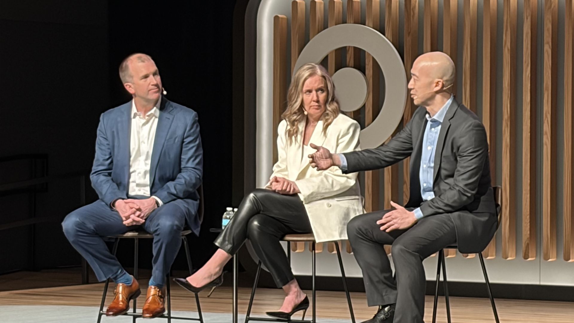 Three people sitting on stools on a stage with wood panel background and a large circular logo. The man on left wears blue suit and tan shoes, woman in middle wears white jacket and black pants, man on right gestures while speaking in dark suit.