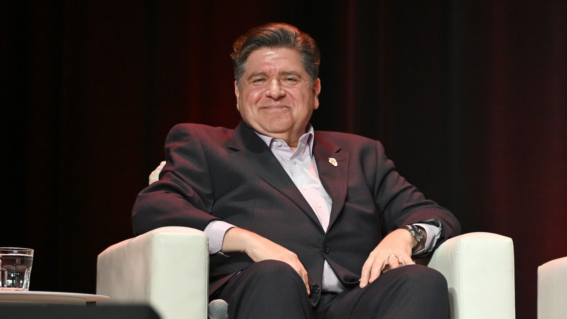 Man in a dark suit sitting on a white chair with a slight smile, against a dark background, with a glass of water on a nearby table.