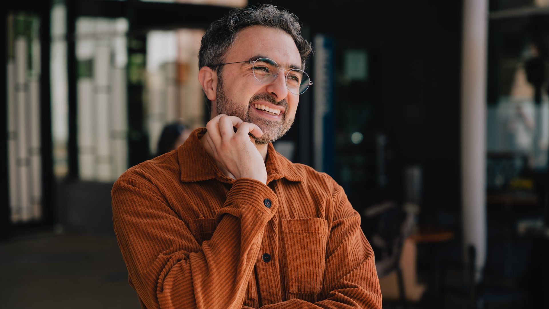 Smiling man with glasses, curly hair, and beard wearing a rust-colored corduroy shirt, standing indoors with crossed arms and a hand touching his neck.