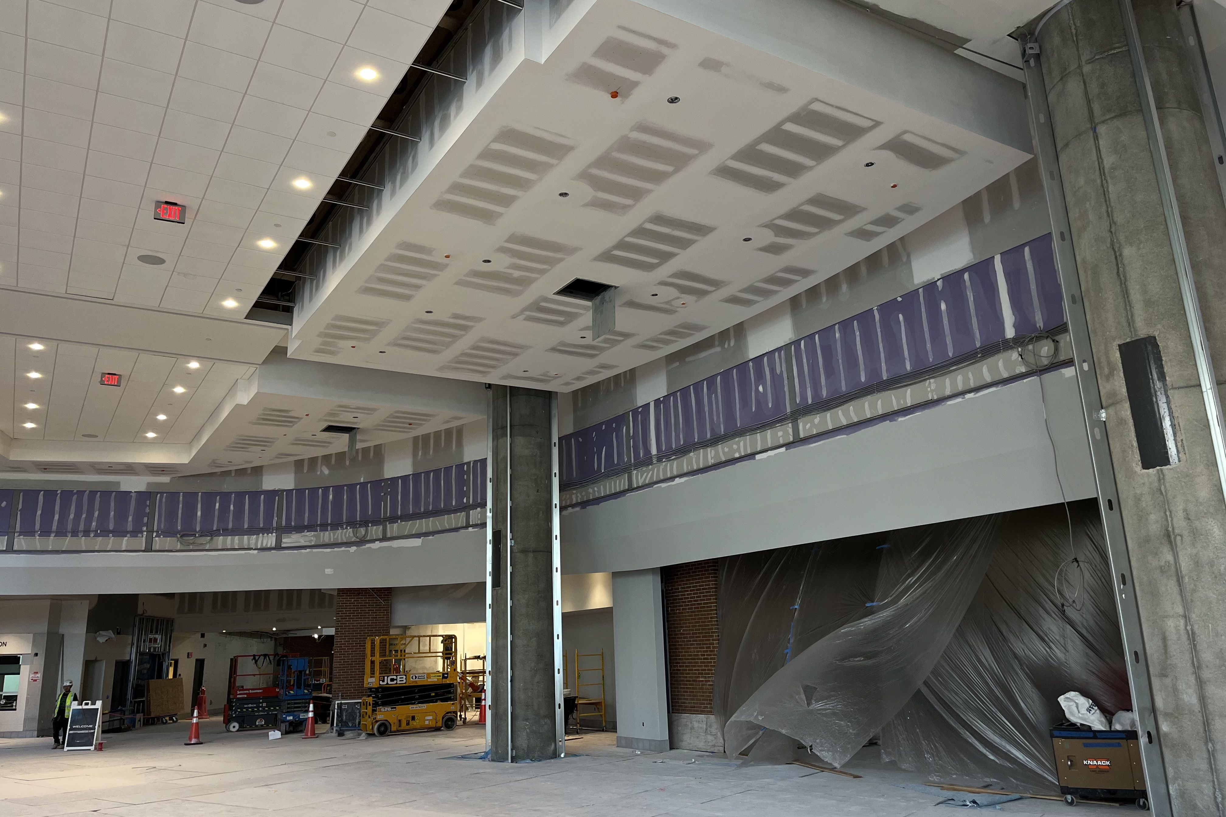 Interior of a large space under construction with exposed drywall, purple drywall panels around upper walls, concrete columns, construction equipment, plastic sheet covering an opening, and workers in safety vests.