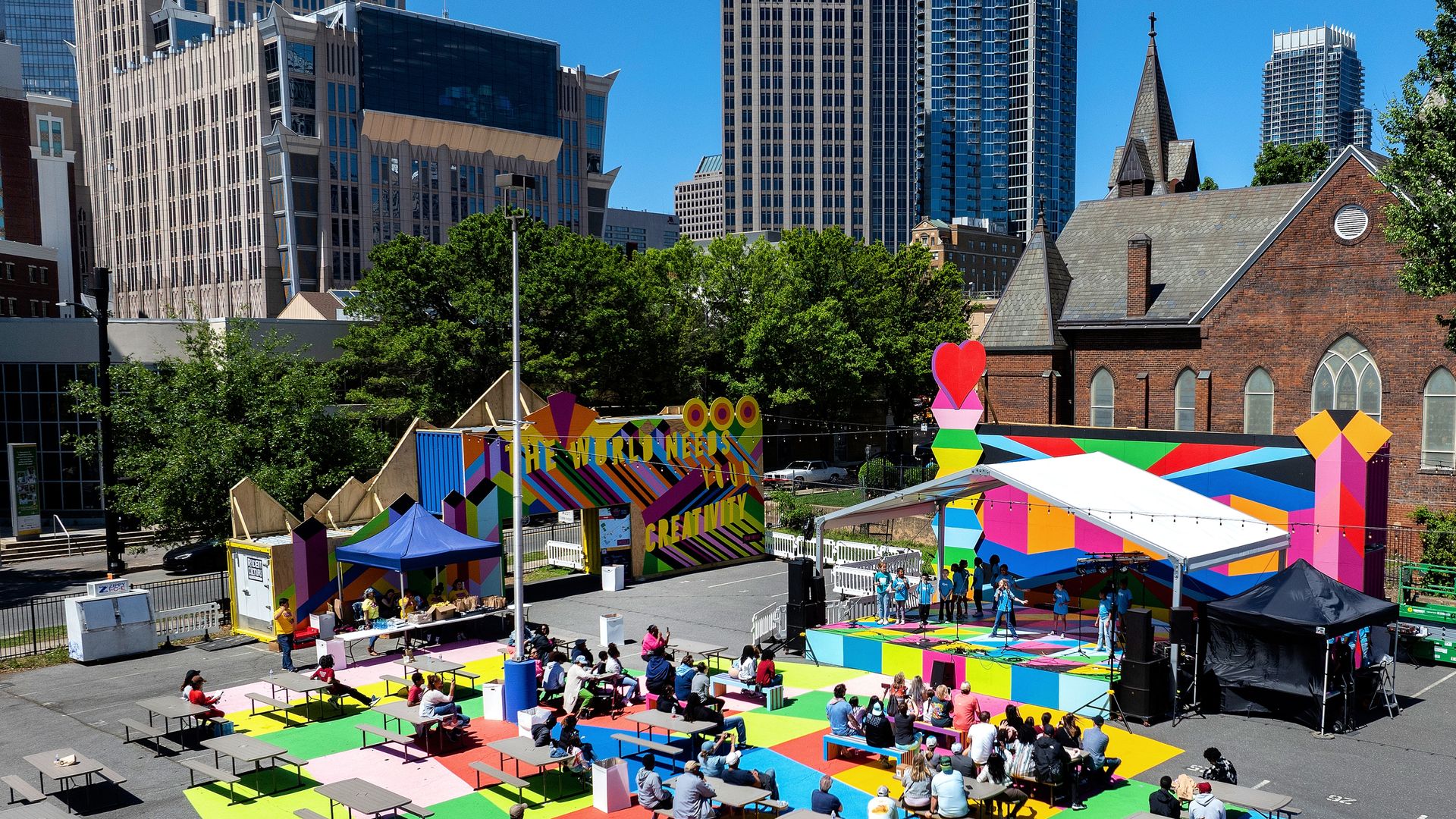 Victoria Yards in Uptown with the city skyline in the background. Someone performs on stage as people sit on colorful spaces and watch. 