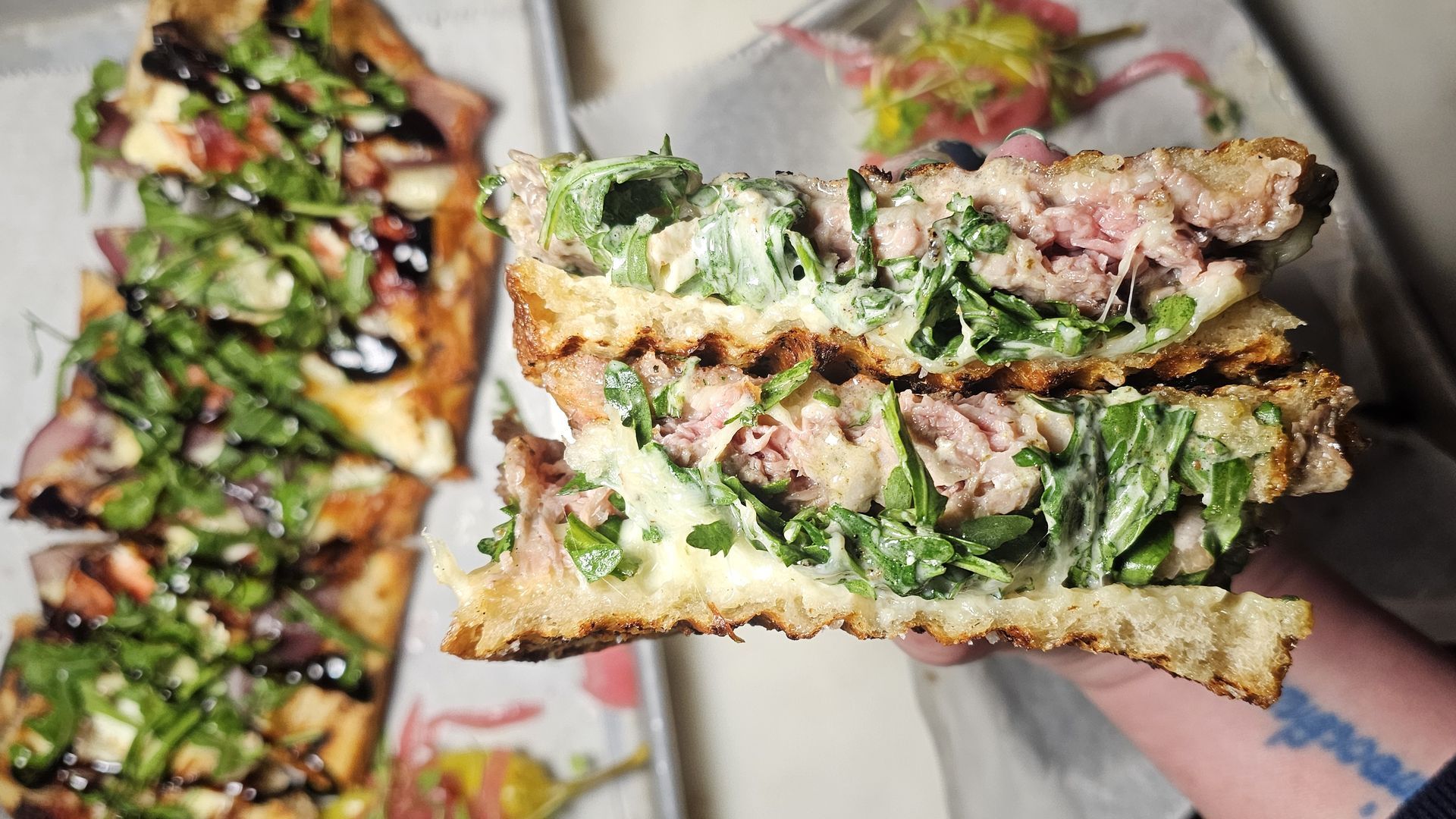 Hand holding a sandwich with toasted bread, melted cheese, leafy greens, and sliced meat, with a flatbread pizza topped with greens and balsamic glaze on a tray in the background.