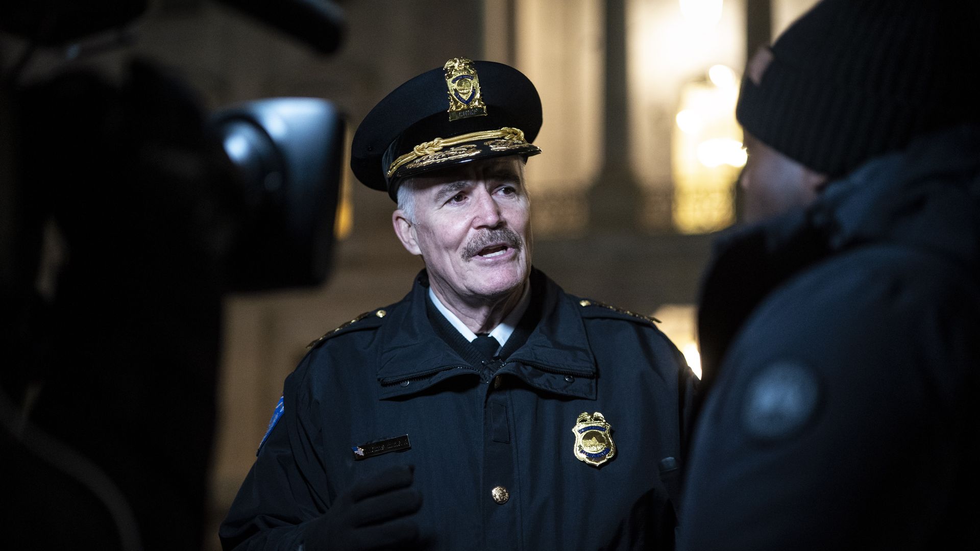 Capitol Police Chief Thomas Manger, wearing a police jacket and hat, stands in front of a camera at the Capitol at night.