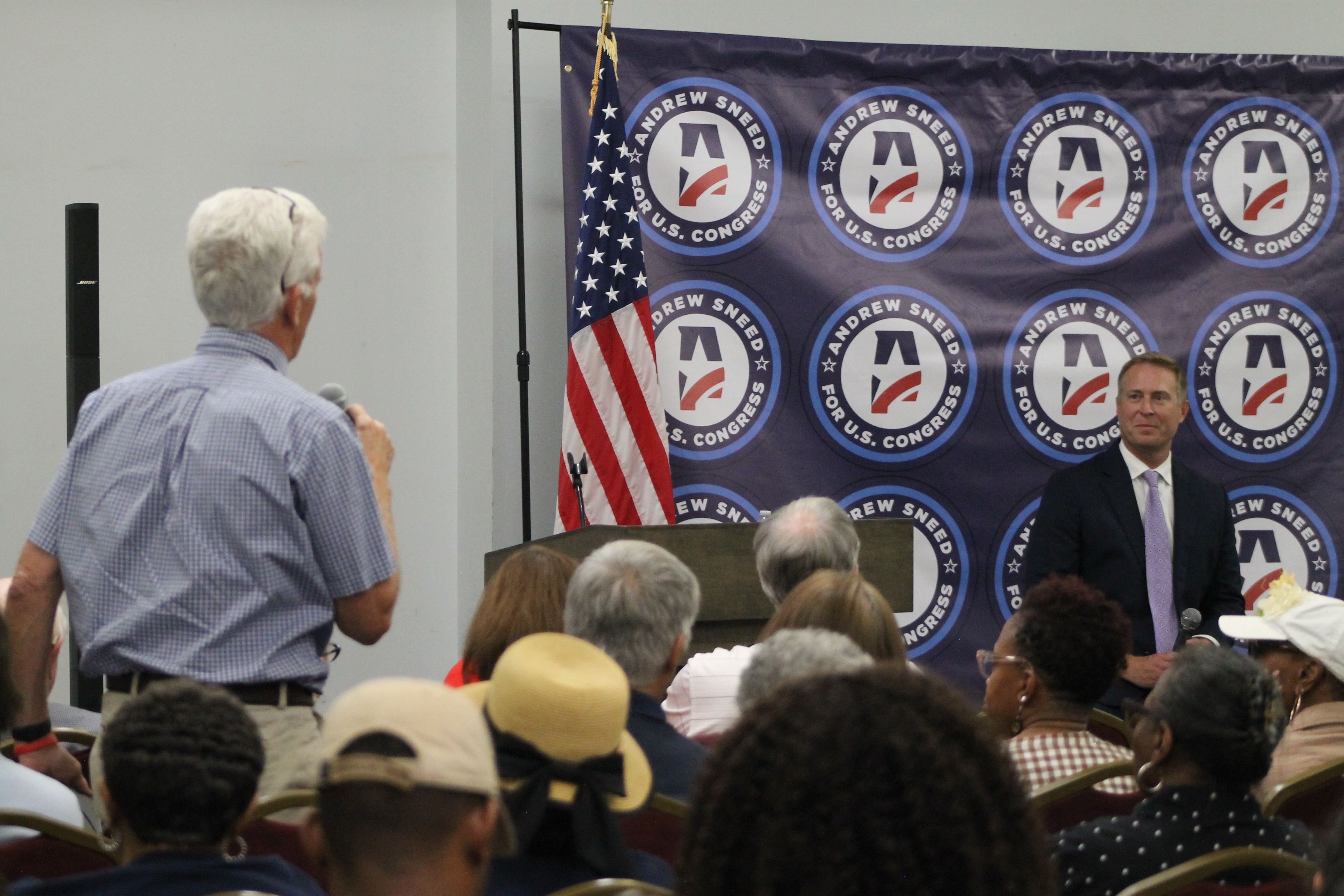 Audience at a political event watches a suited speaker at a podium; a dark blue backdrop features circular banners reading Andrew Sneed for U.S. Congress with blue borders and an American flag nearby.