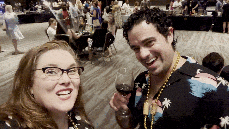 Image shows Carlie and Kenny Lopez smiling and waving a glass of red wine in front of the camera.