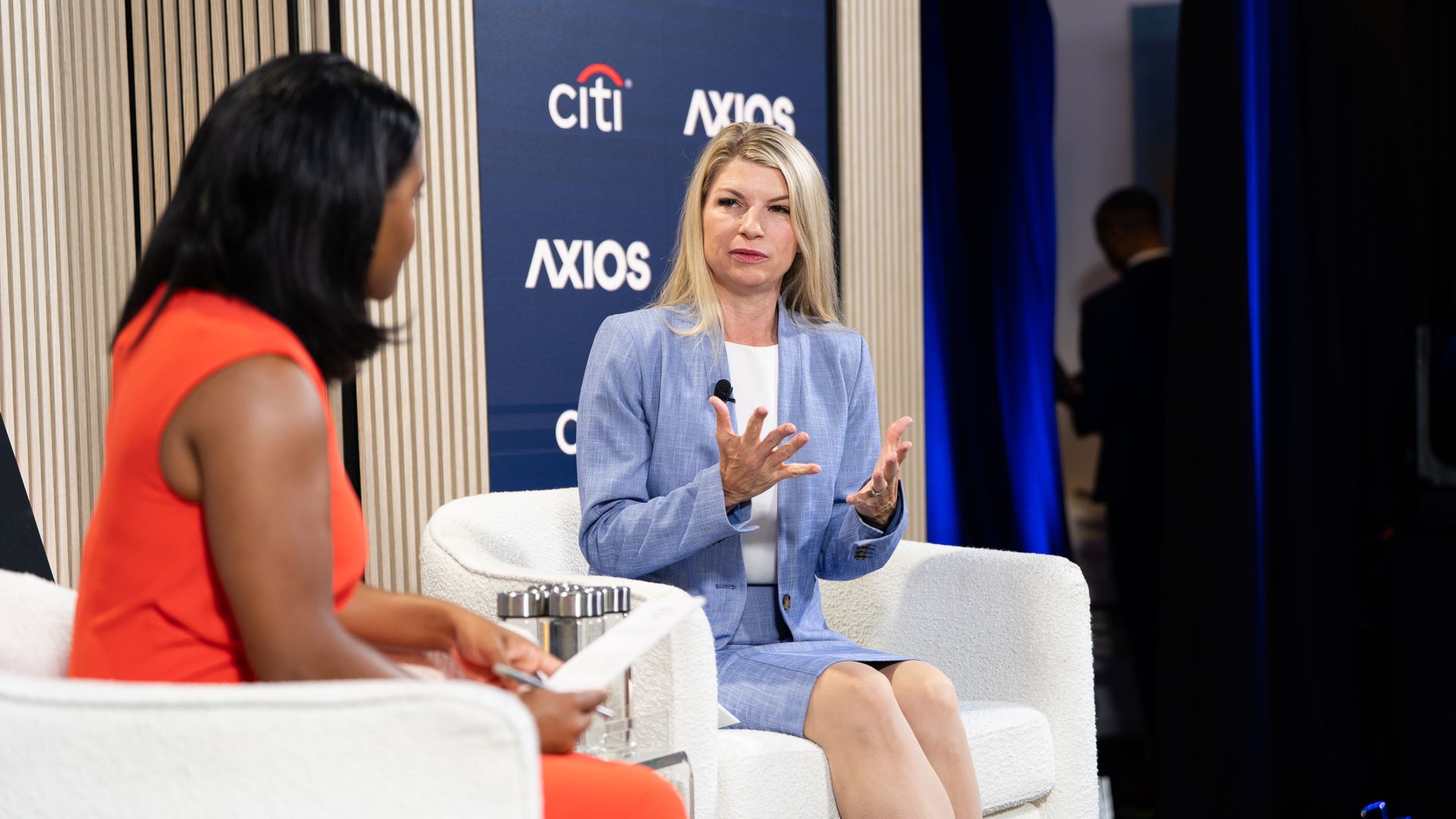 Panel discussion: a blonde woman in a light blue blazer speaks and gesticulates, facing a dark-haired interviewer in an orange dress. White chairs, blue Axios and Citi backdrop.