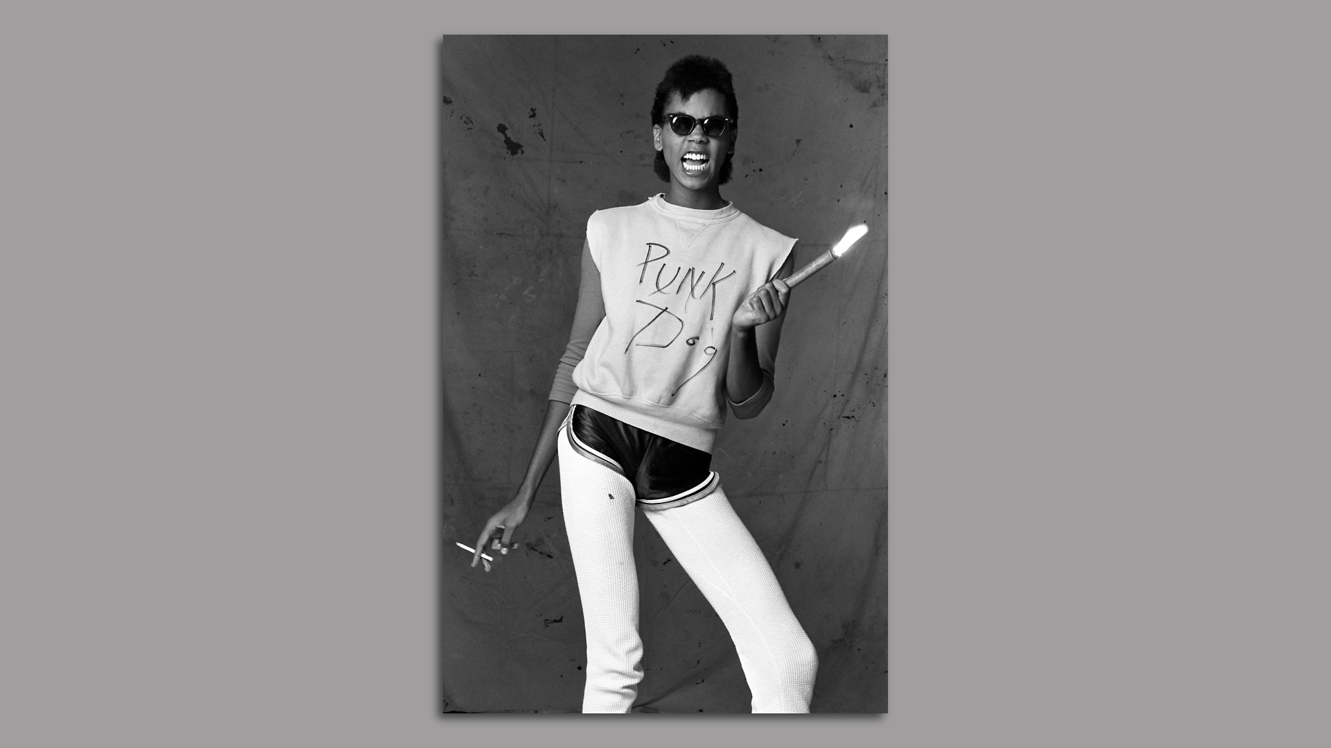 A black and white photo of a young RuPaul wearing sunglasses and a shirt saying "punk dog" while holding a flare and cigarette