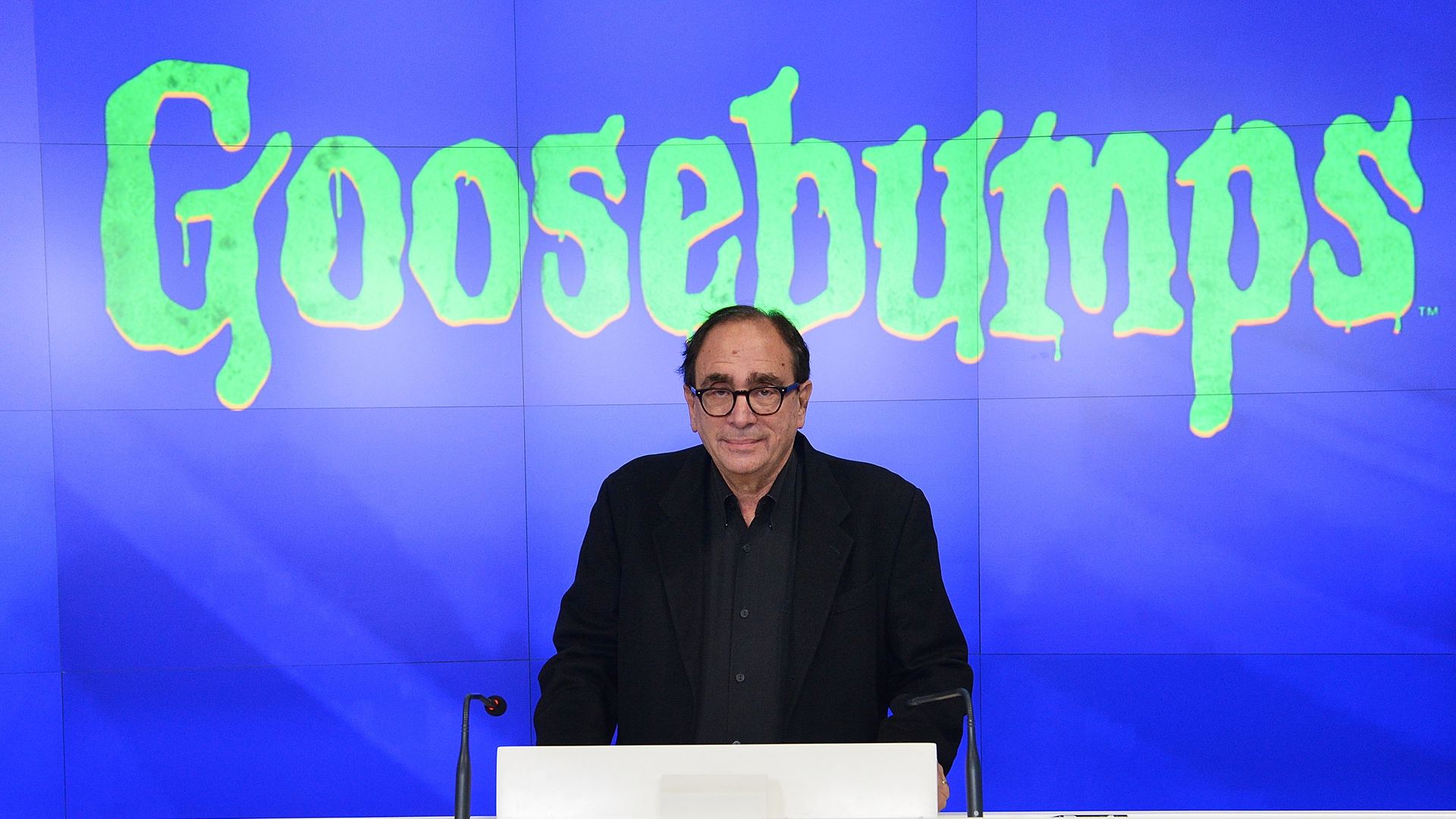 Man with glasses in a black blazer stands at a white Nasdaq podium, speaking before a blue backdrop featuring a large neon-green "Goosebumps" logo.