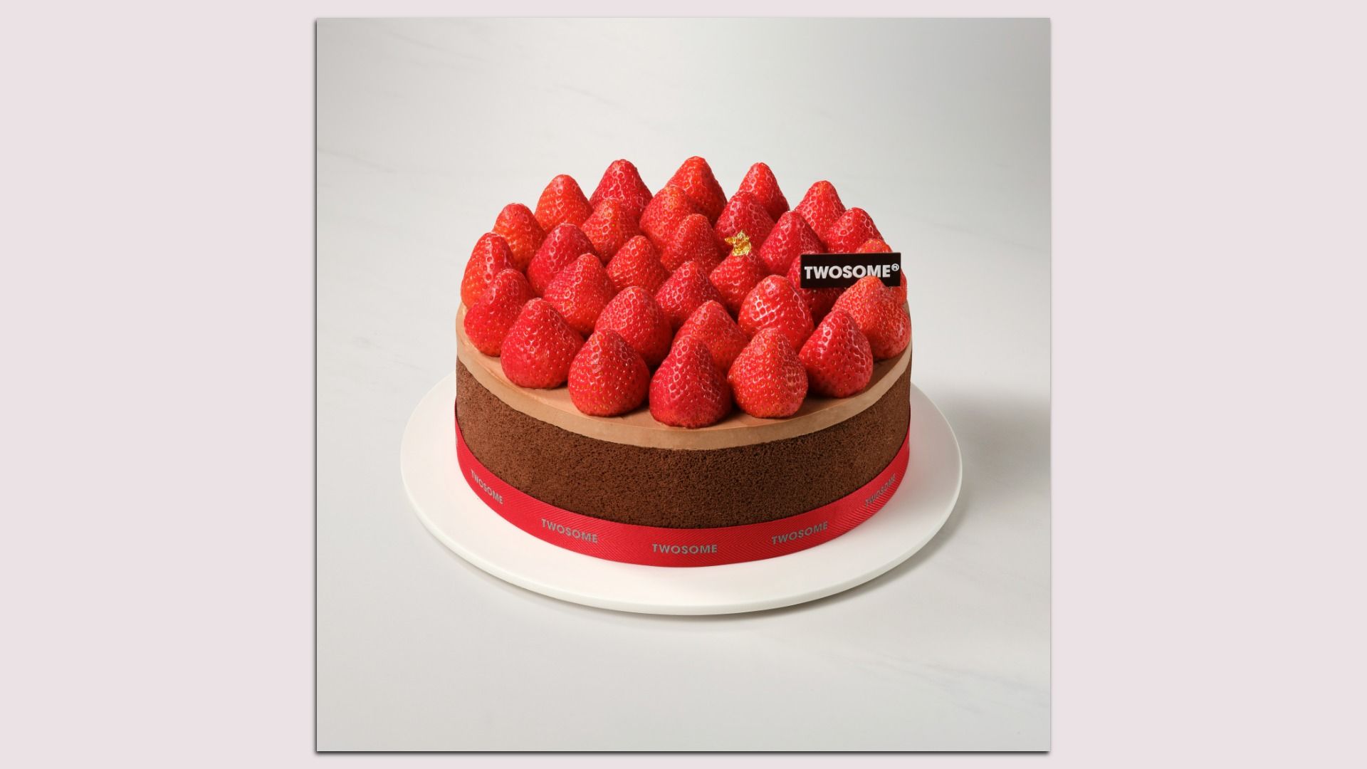 Chocolate cake topped with a pyramid of bright red strawberries; a black TWOSOME label sits among the fruit, with a red ribbon around the base on a white plate against a pale backdrop.