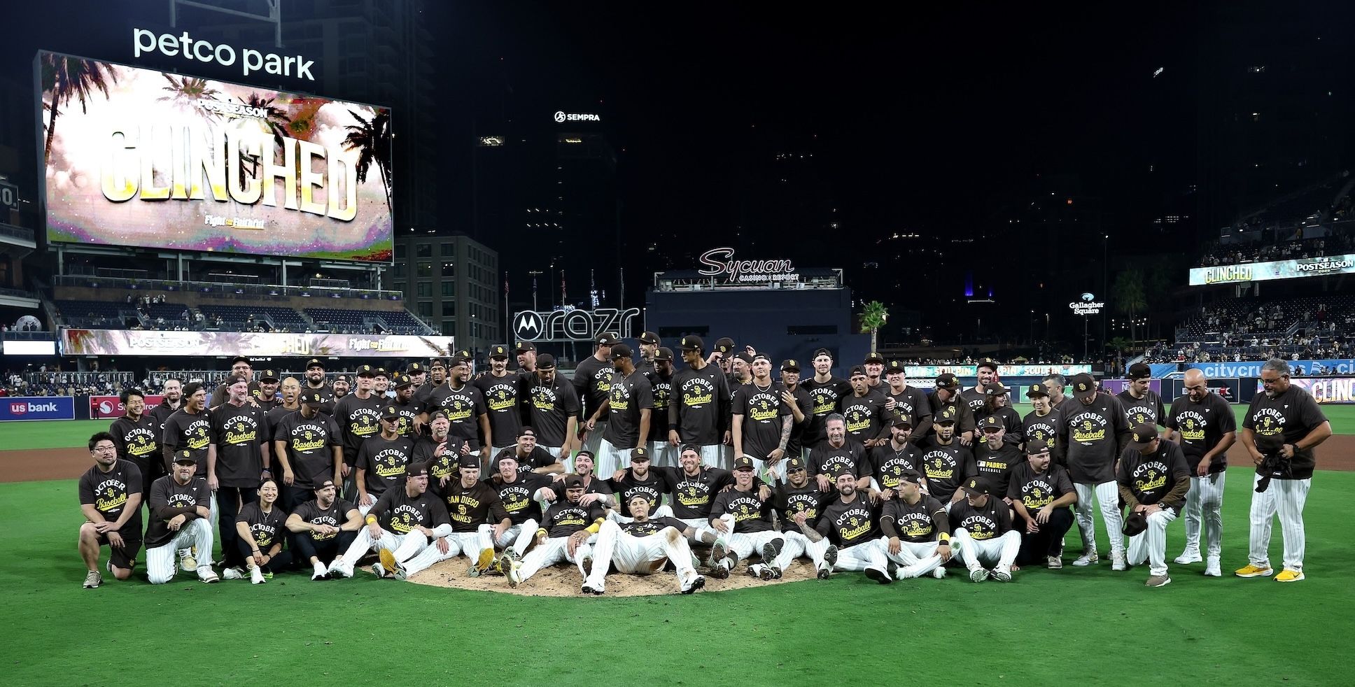 Padres players and staff pose for a group photo on the field wearing brown shirts that say "October Baseball."