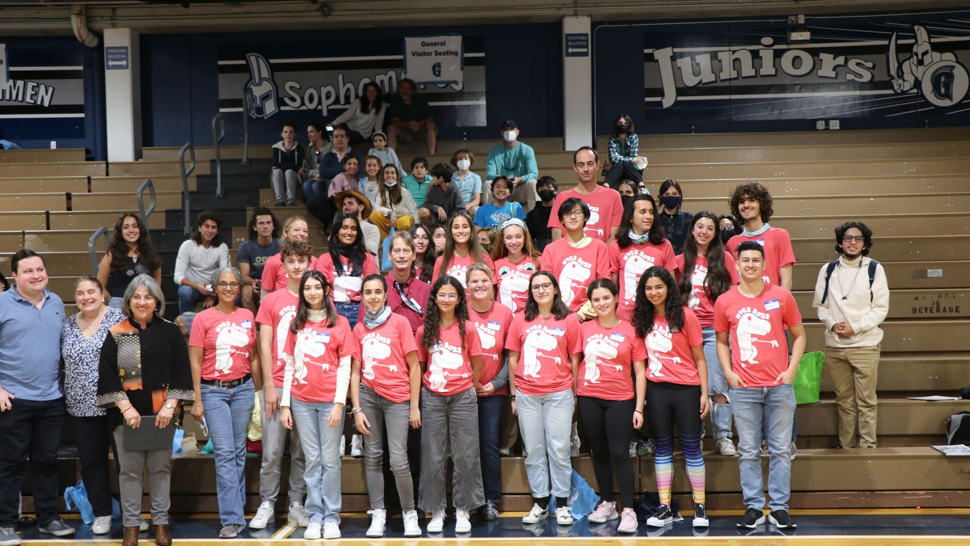 A group of teens wearing pink t-shirts poses in front of wooden school bleachers.