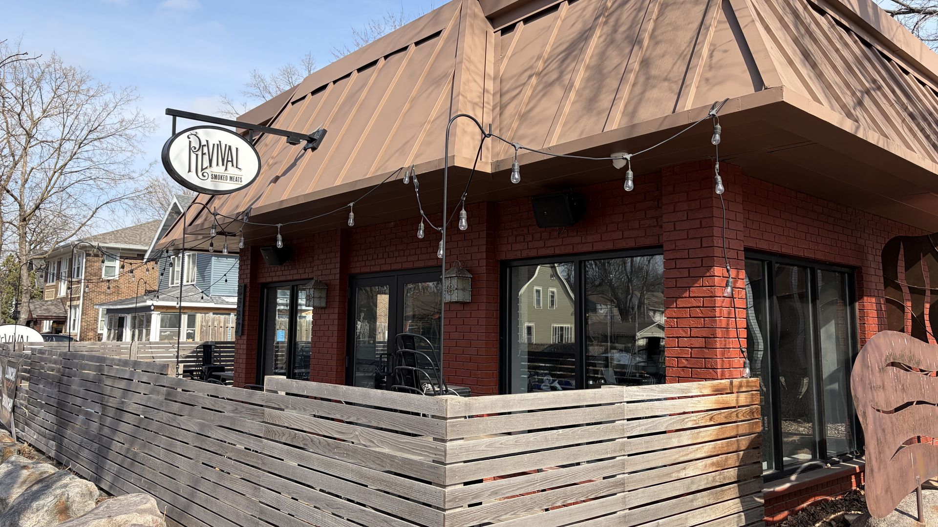 Red brick building with brown metal roof and wooden fenced patio featuring string lights; sign reads "Revival Smoked Meats" on a clear day with leafless trees nearby.