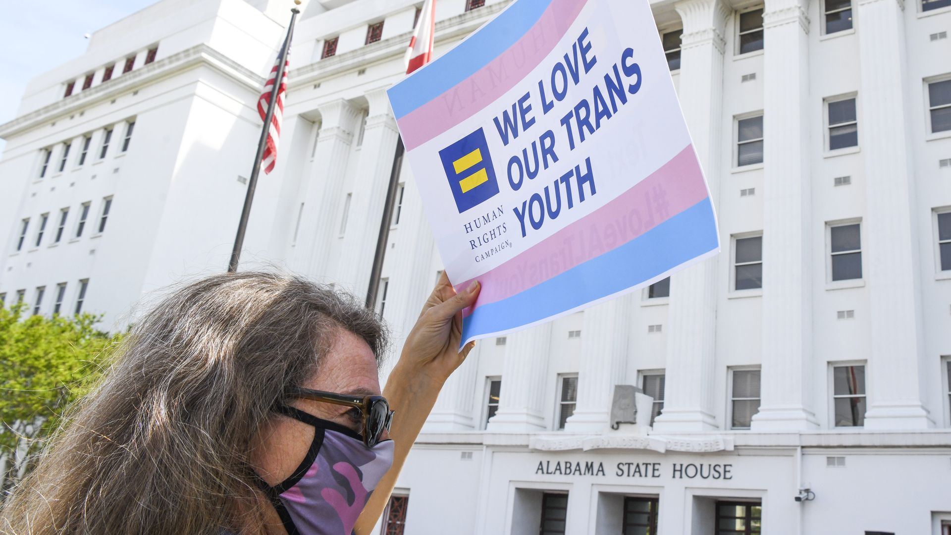 Photo of a person holding a sign that says "We love our trans youth" with the trans flag colors outside the white Alabama statehouse