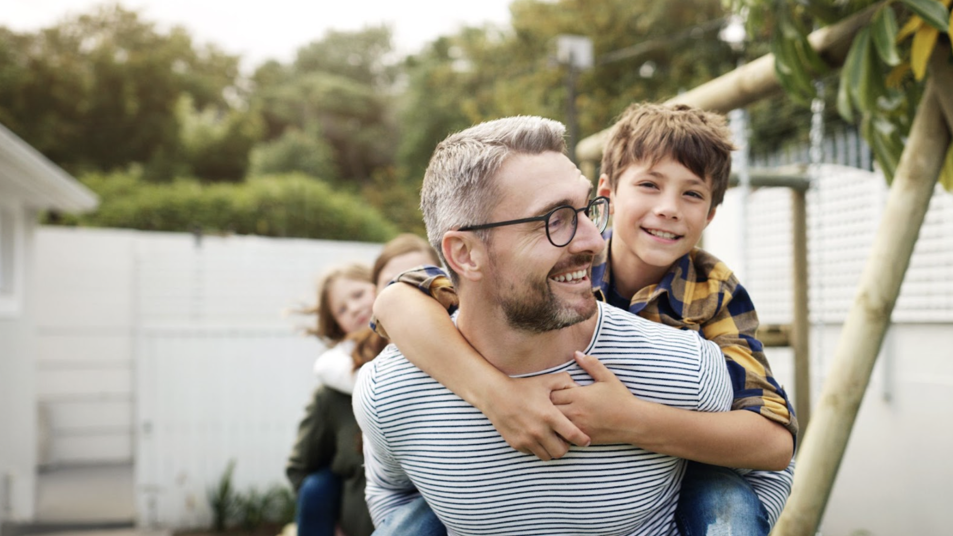 Smiling father with gray hair and glasses wearing a white striped shirt carries his young son in a yellow and blue plaid shirt on his back outdoors near a white fence and greenery.