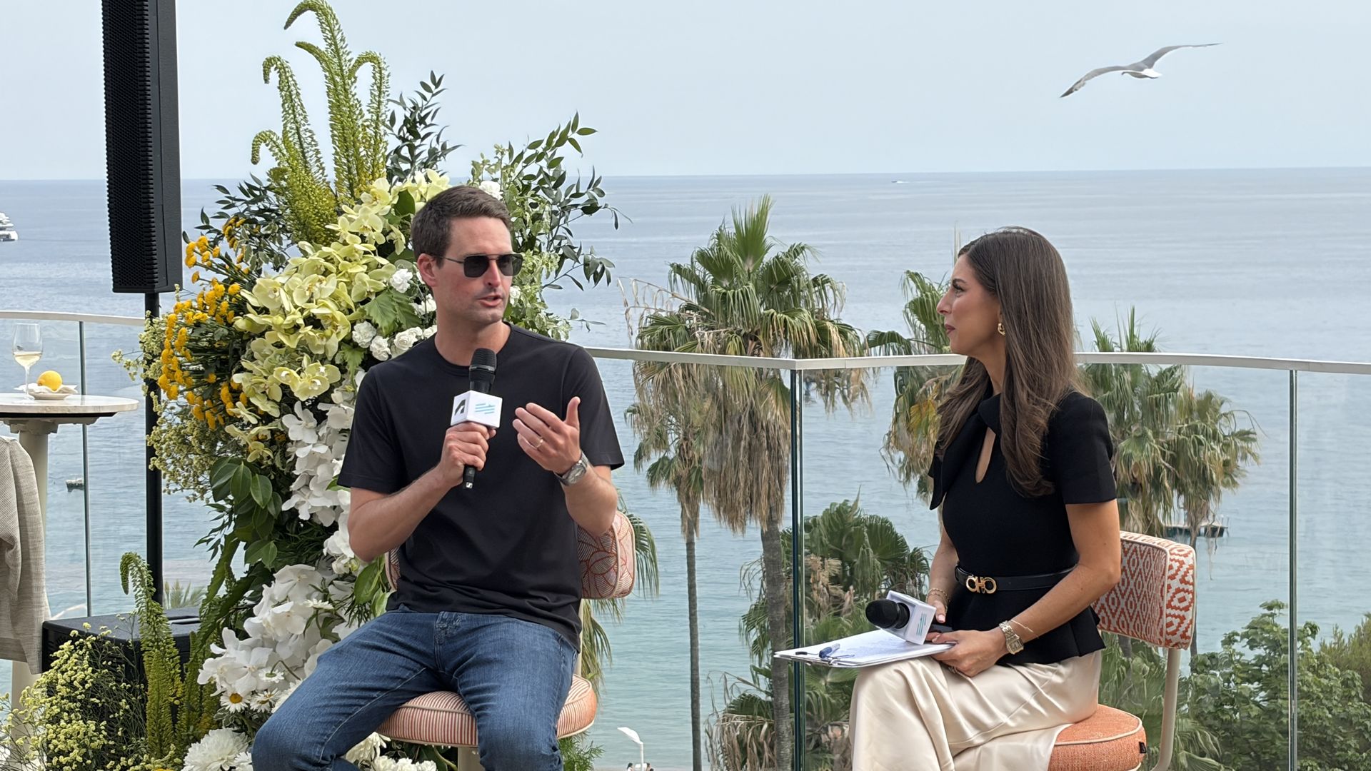 Snap CEO Evan Spiegel speaks to Axios' Sara Fischer by the sea; Spiegel in a black shirt and sunglasses speaks into a microphone while Fischer in black and beige holds notes. Floral arrangements, palms, and a seagull over the ocean.