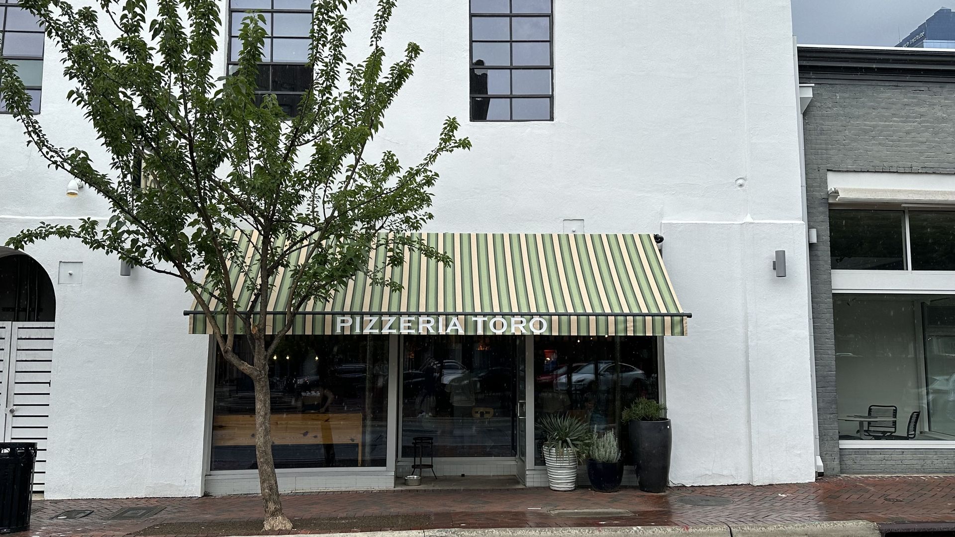 Front of Pizzeria Toro with green and beige striped awning, white building facade, tree in front, and potted plants near entrance on rainy sidewalk.