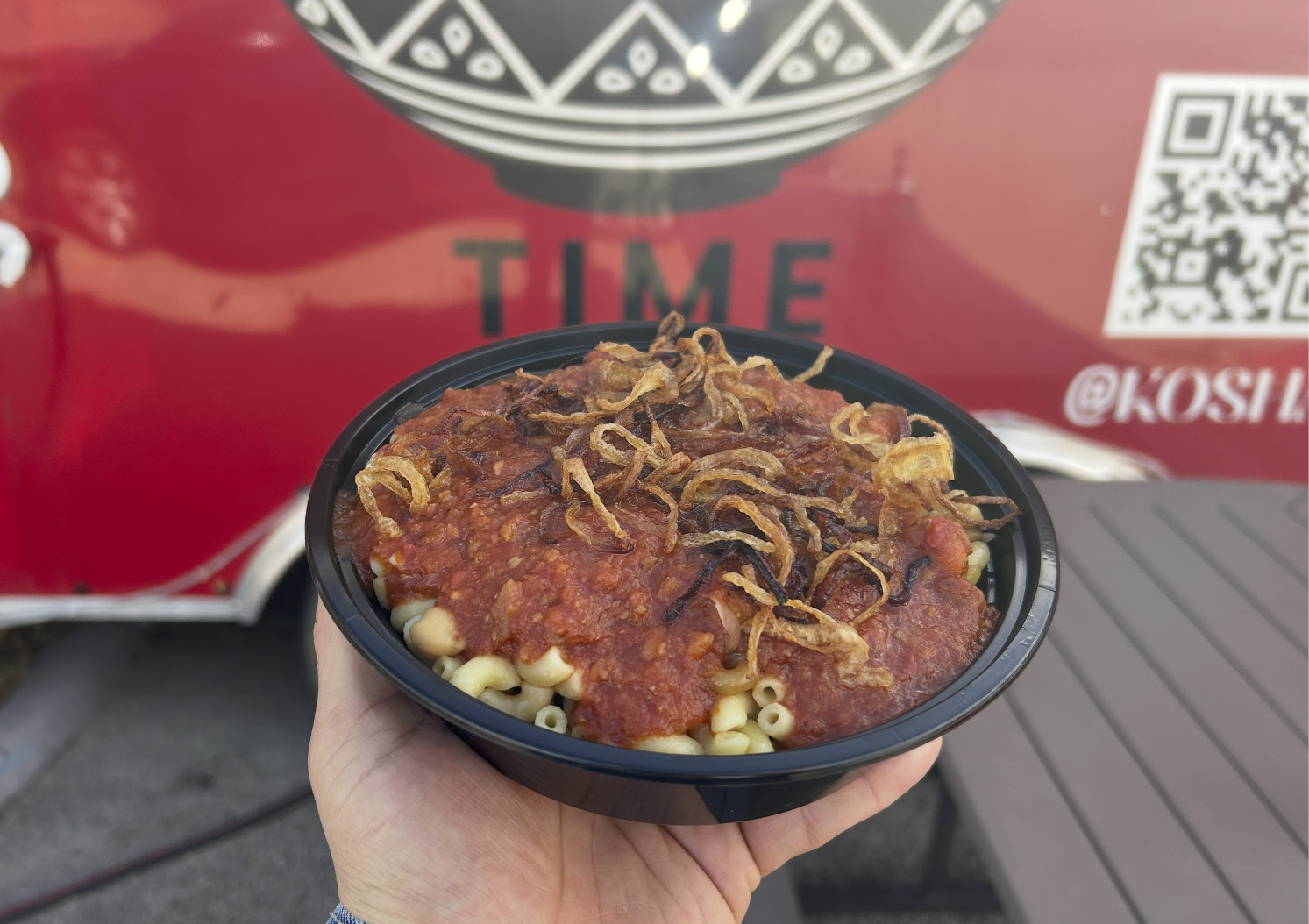 A bowl of Koshary, featuring rice, lentils, pasta, chickpeas and crispy fried onions, topped with a rich tomato sauce. The bowl is held in hand in front of the Koshary Time food truck. 