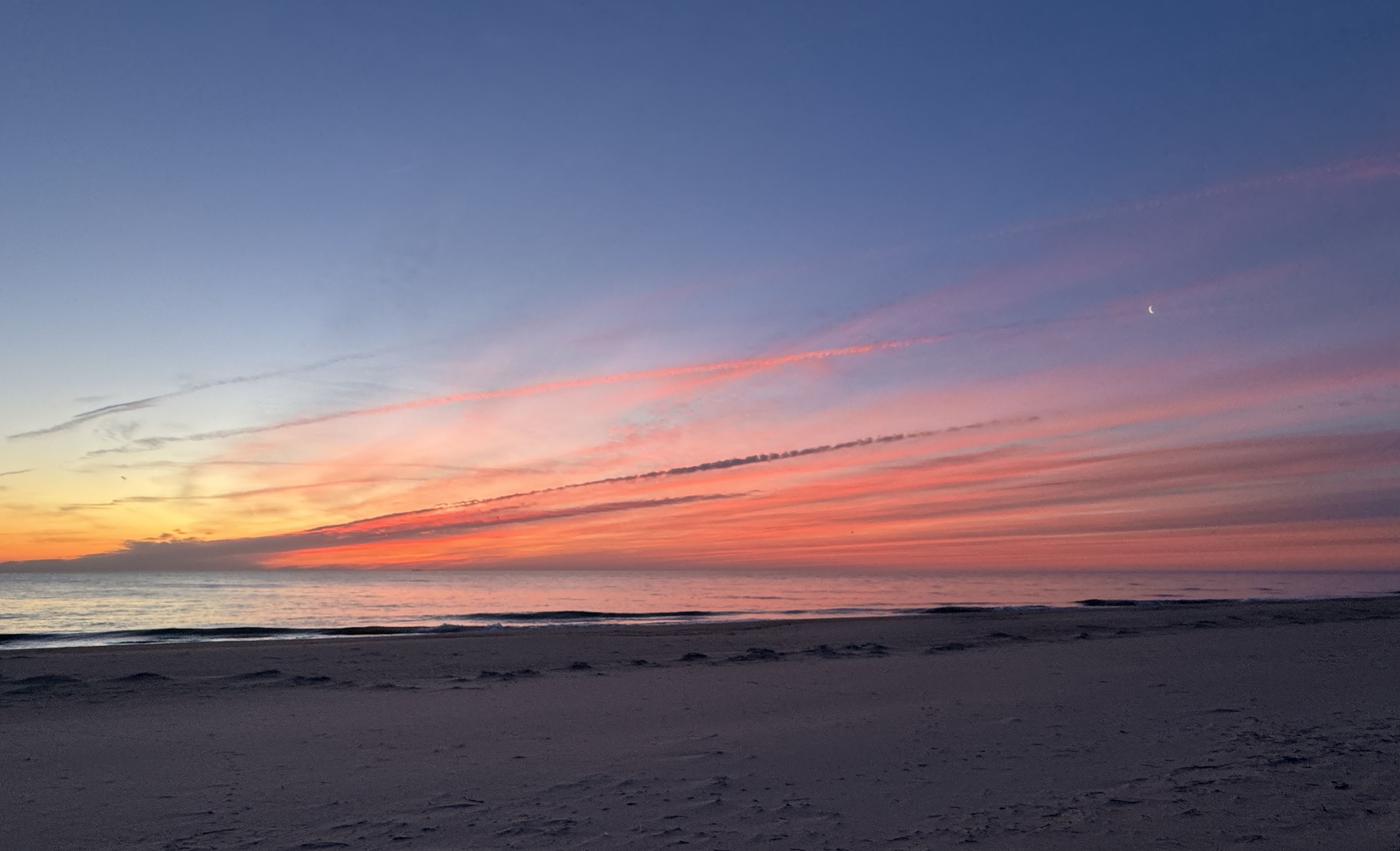 Serene beach at sunset with a blue-to-pink sky, orange clouds, calm sea on the horizon, and a sandy foreground; a small crescent moon appears in the upper right.