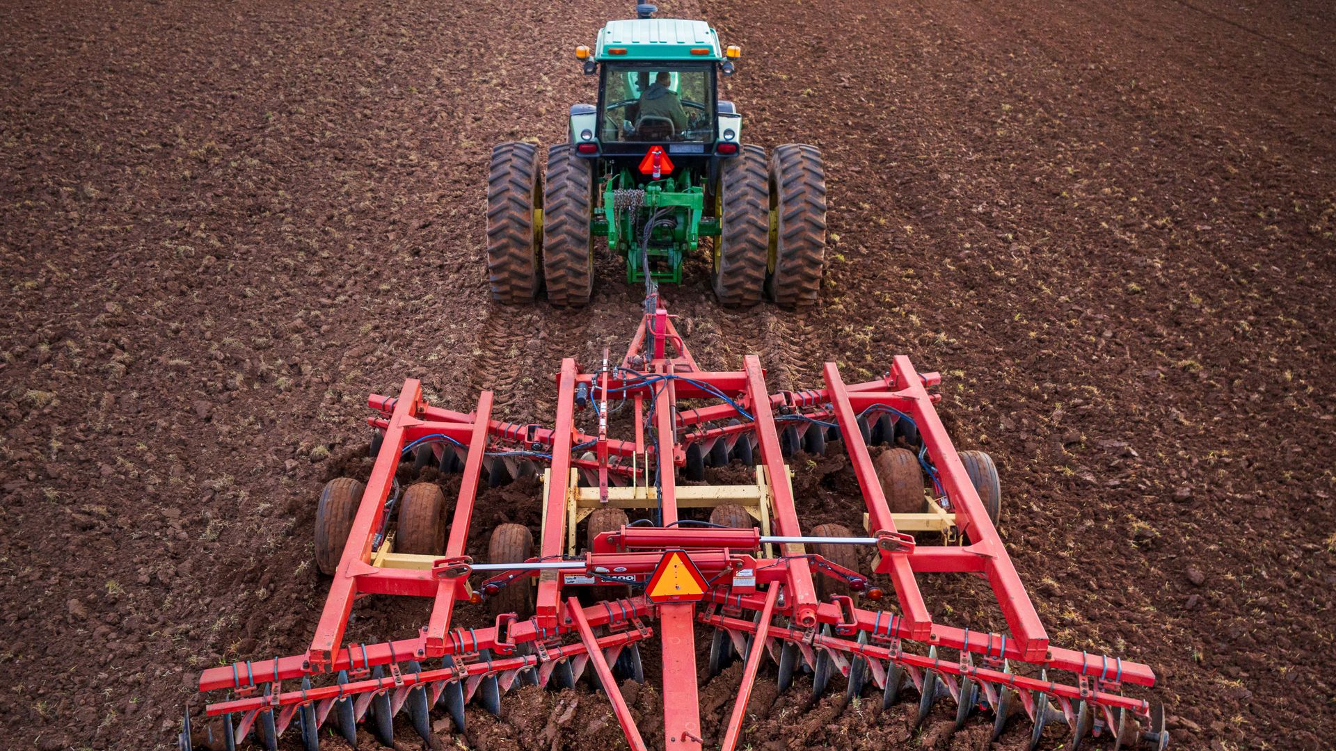Top-down view of a green tractor pulling a red cultivator across a brown, freshly tilled field; oversized rear tires, orange slow-moving sign, and blue hoses visible.