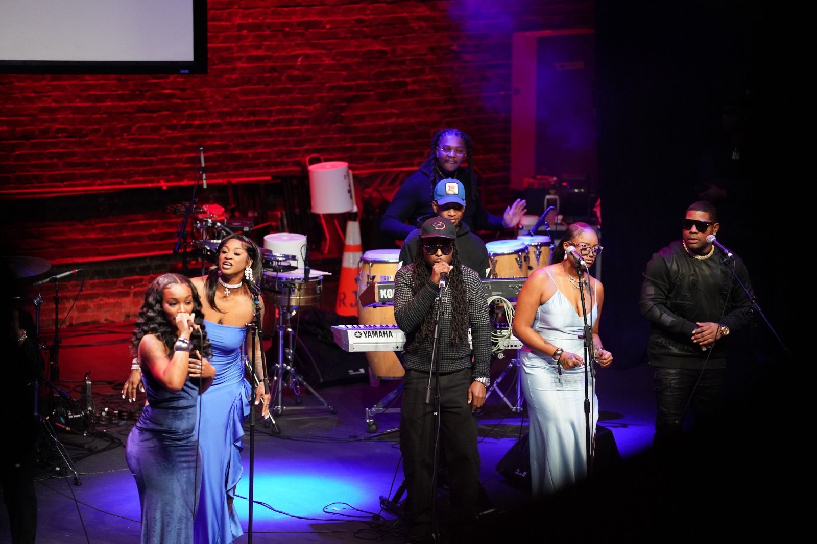 Six musicians on stage performing with red brick wall backdrop; three women in blue dresses singing, two men in black with sunglasses singing, one man playing percussion in background.