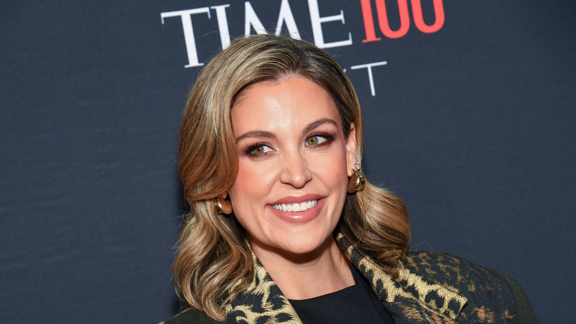 Woman with wavy blonde hair and gold earrings smiling, wearing black top and leopard print jacket, standing in front of a backdrop with "TIME 100" text in white and red.