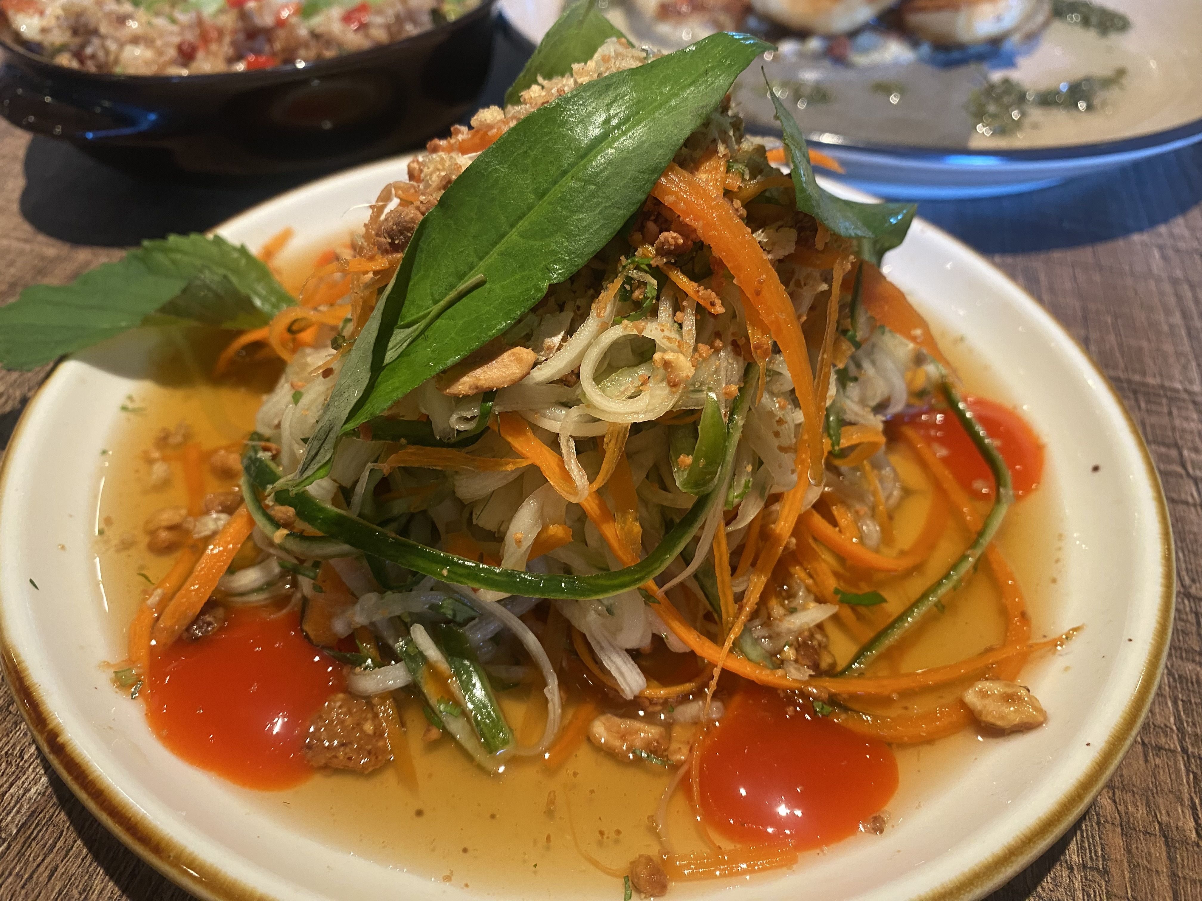 A plate of colorful fresh salad with shredded carrots, green herbs, peanuts, and red chili sauce on a white plate with a wooden table background.