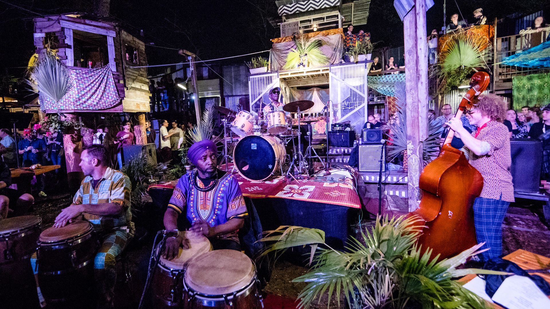 Drummers and an upright bassist perform at the Music Box Village. They are surrounded by eclectic tiny houses and an audience.