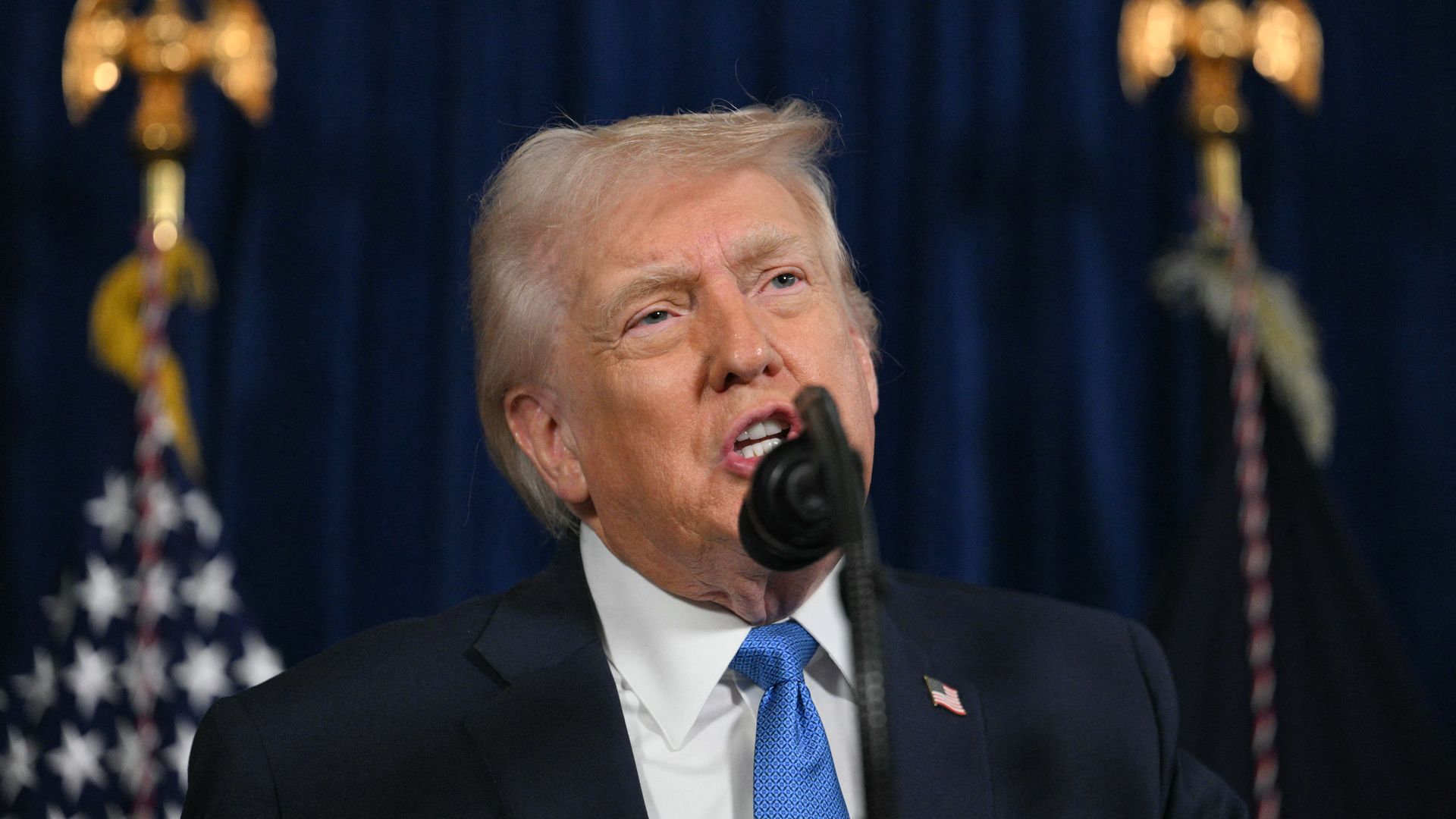 Close-up of a man with light blond hair in a dark suit and blue tie speaking into a microphone, with blurred American flags and gold eagle finials in the background.