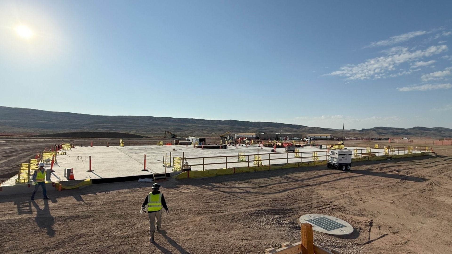 Construction site on TerraPower's first nuclear plant shows a desert area with concrete foundation, two workers in yellow vests and helmets, surrounded by safety rails and orange cones under a clear blue sky with bright sun.