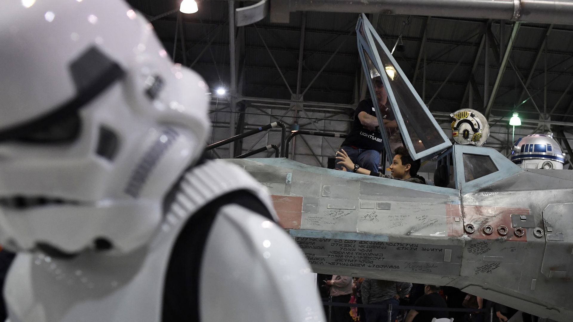 In the foreground, a stormtrooper characters from Star Wars tilts their head, while in the background, a person sitting inside the cockpit of a replica starship takes a photo, while another person stands next to them.