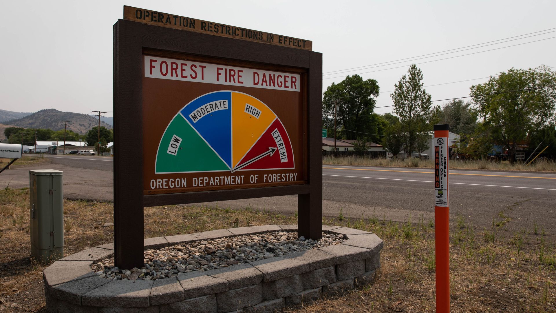 Roadside sign by the Oregon Department of Forestry displaying "Forest Fire Danger" with a circular risk gauge from Low to Extreme; brown frame, rocky base, rural setting with hills and power lines.