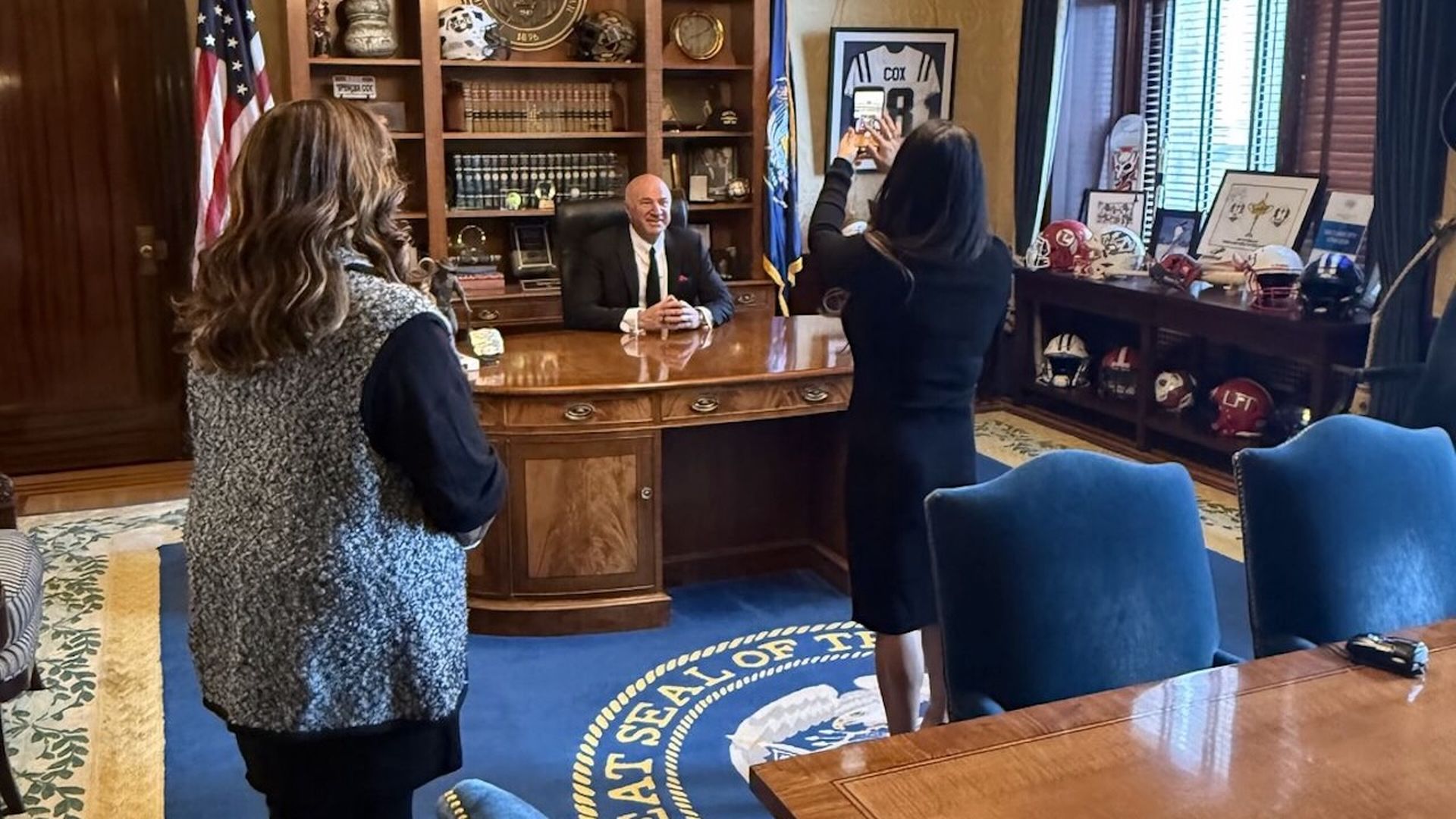 Kevin O'Leary sits at the Utah Governor's desk while a woman takes his picture and another woman looks on.