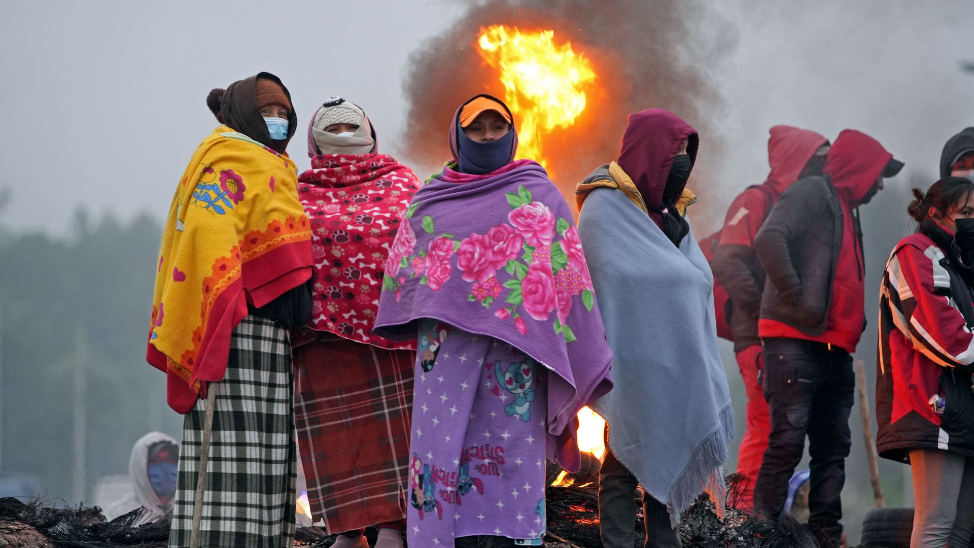 A group of women covered in blankets, masks and hoods stand in front of a large blaze during protest in Peru 