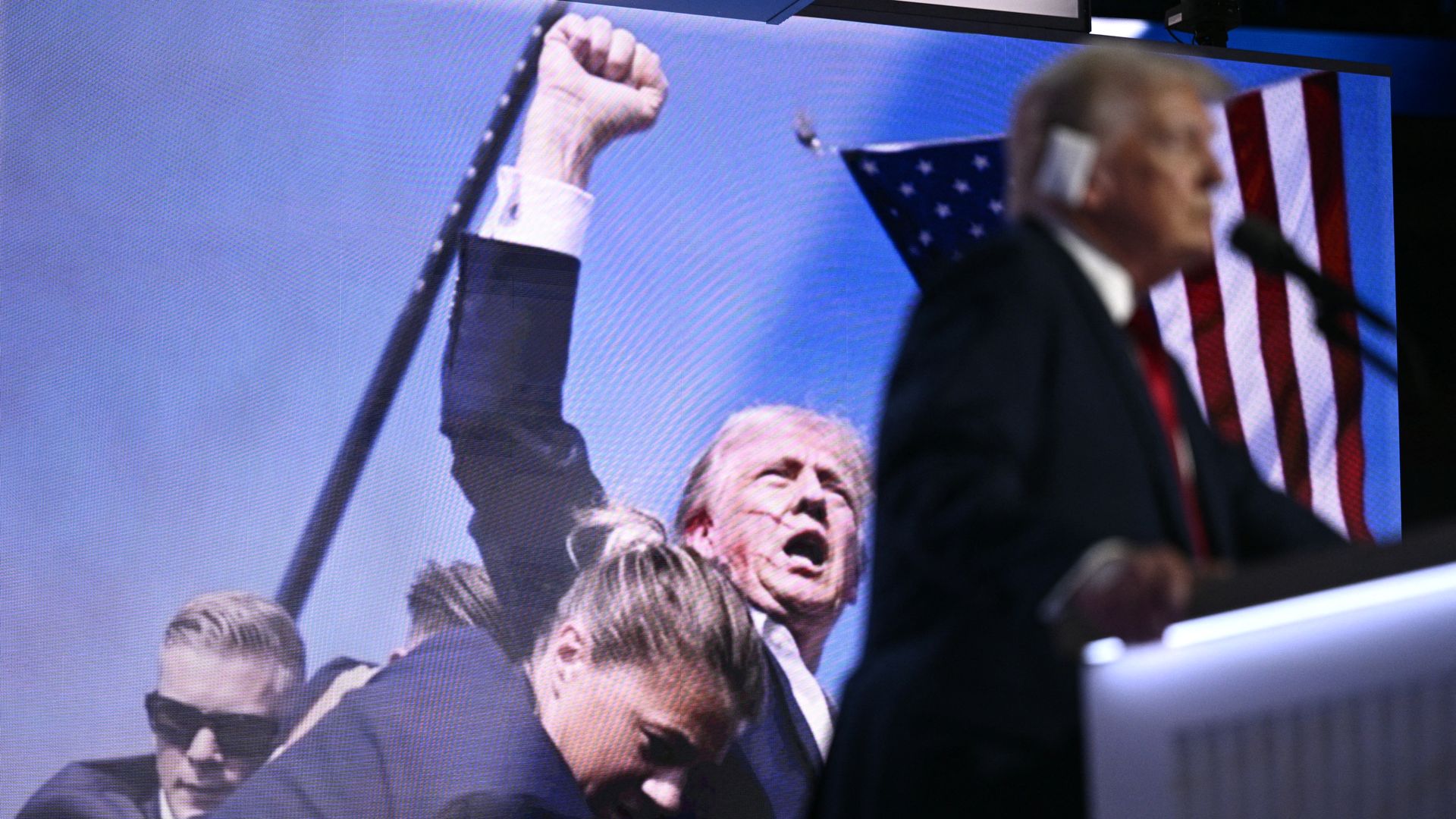 Donald Trump speaks at a convention podium while a large screen behind him shows an image of him raising his fist after an assassination attempt.