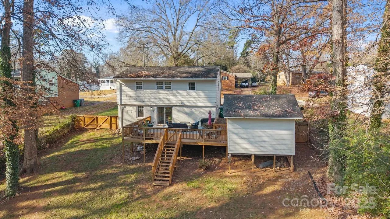 Back of a two-story beige house with a raised wooden deck and stairs, next to a small detached shed. Trees with brown and few leaves surround the yard under a blue sky.