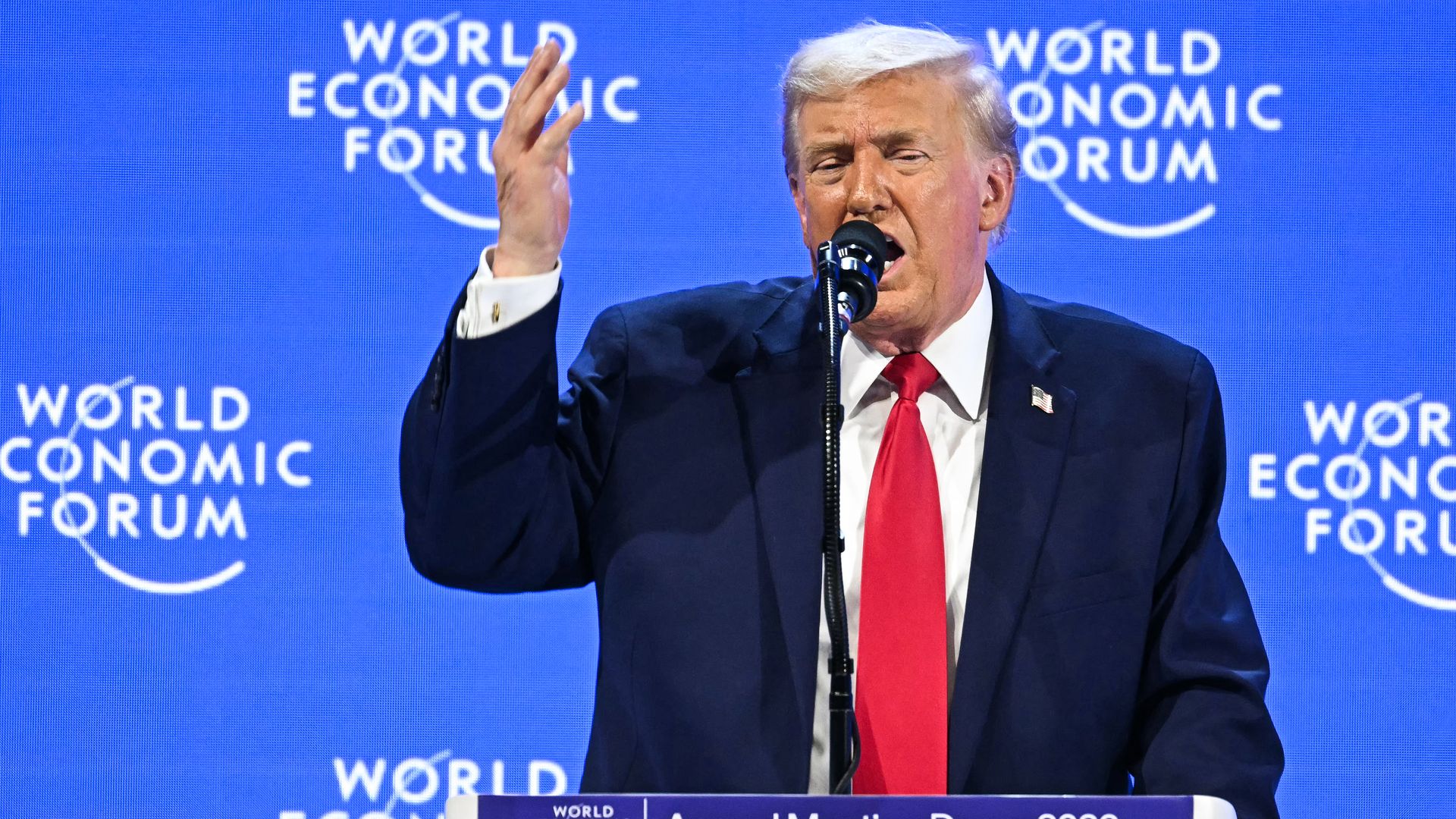 Donald Trump standing at a podium gesturing with his right hand while speaking at the World Economic Forum in Davos, with a blurred blue background behind him.
