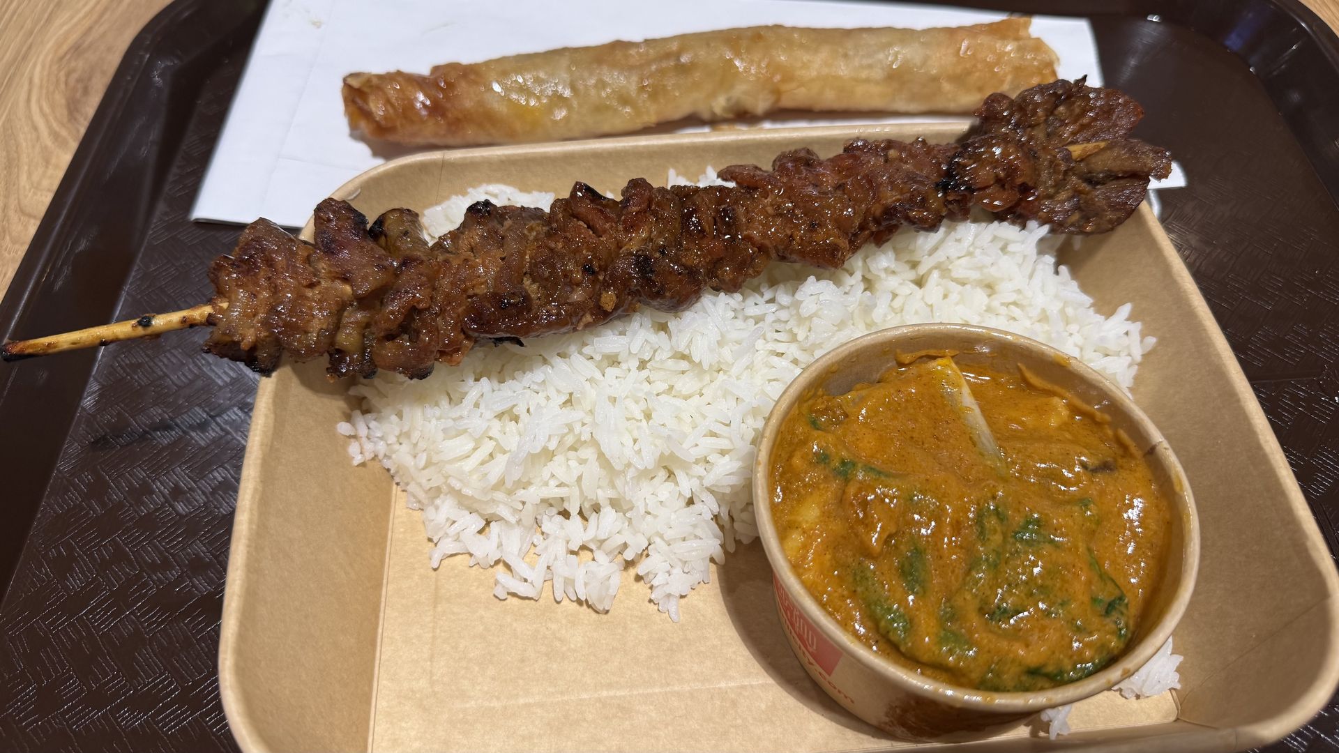 Cardboard takeout box with white rice topped by a grilled pork skewer covered in sauce, a small cup of brownish orange stew with greens, and a long fried pastry in the background on piece of white wax paper.
