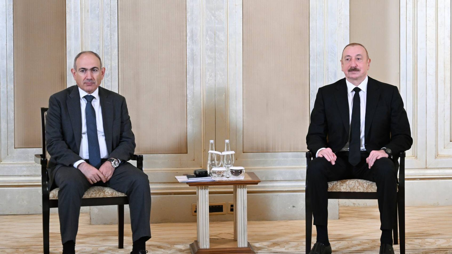 Two men in suits sit on chairs with a small table between them holding two water bottles and glasses, against a beige and white paneled wall background.