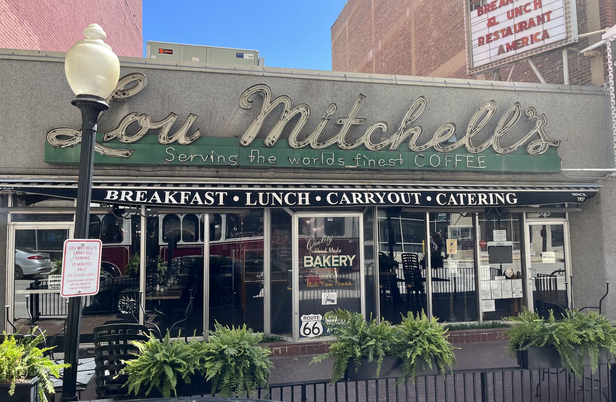 Neon Lou Mitchell's sign on a gray tiled storefront with a green banner reading 'Serving the world's finest COFFEE', black awning 'BREAKFAST LUNCH CARRYOUT CATERING', and potted ferns outside.