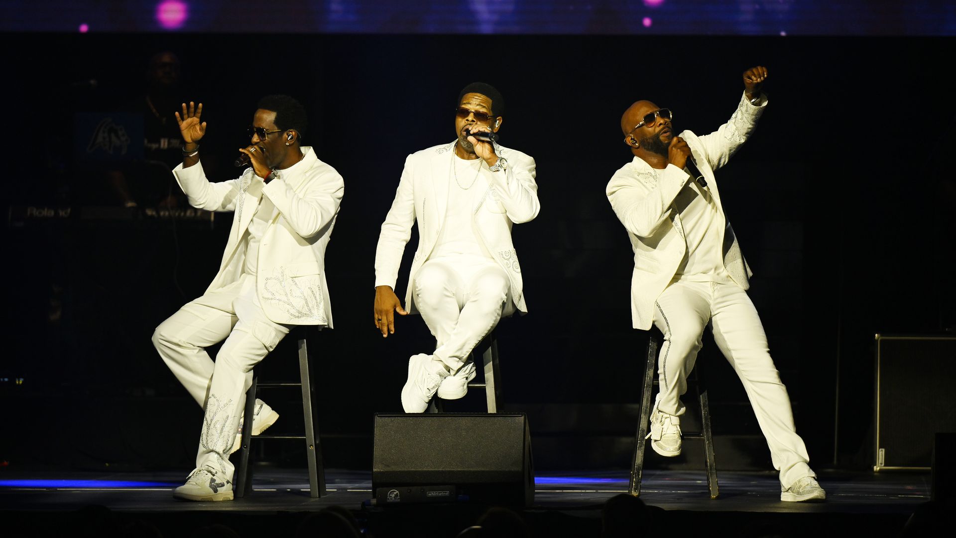 Photo shows three men, dressed in white suits, while singing on stage.