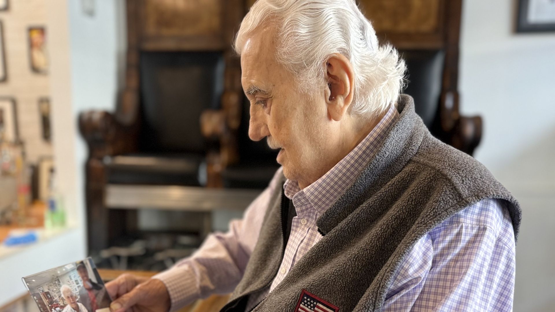 An elderly man with white hair, wearing a plaid shirt and a gray fleece vest with an American flag patch, looks at a photo album at a counter in a cozy shop.